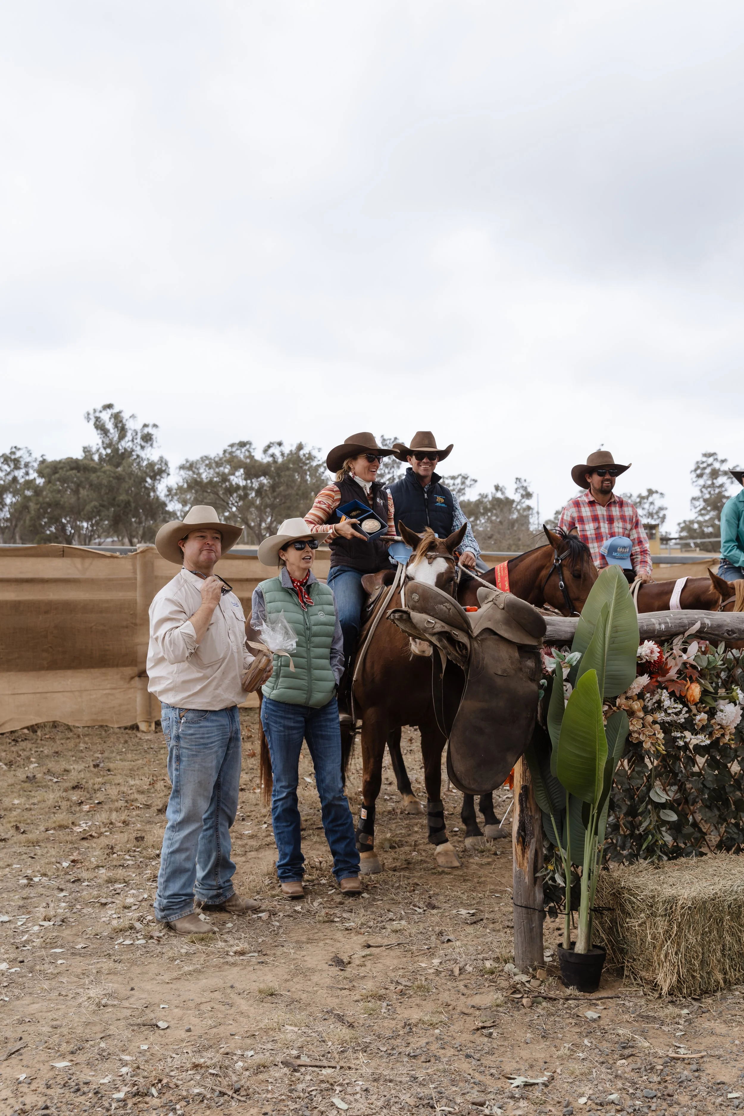 A group of people wearing cowboy hats at an outdoor event, some on horseback and some standing nearby, with a flower arrangement in the foreground.