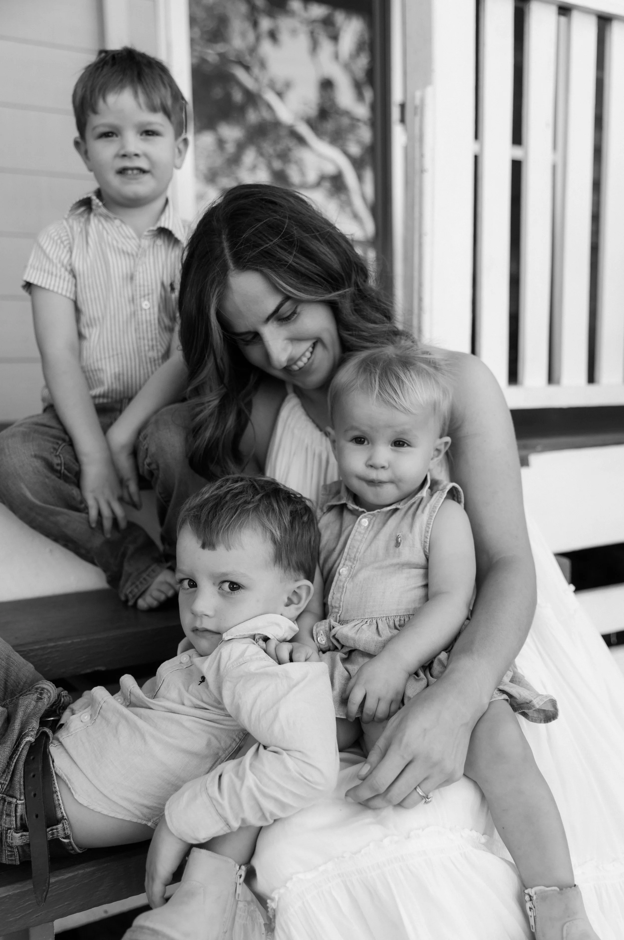 A woman surrounded by five young children, sitting on a porch with a wooden railing and a tree mural in the background. The children are of different ages, and they are all looking at the camera or engaging with the woman.