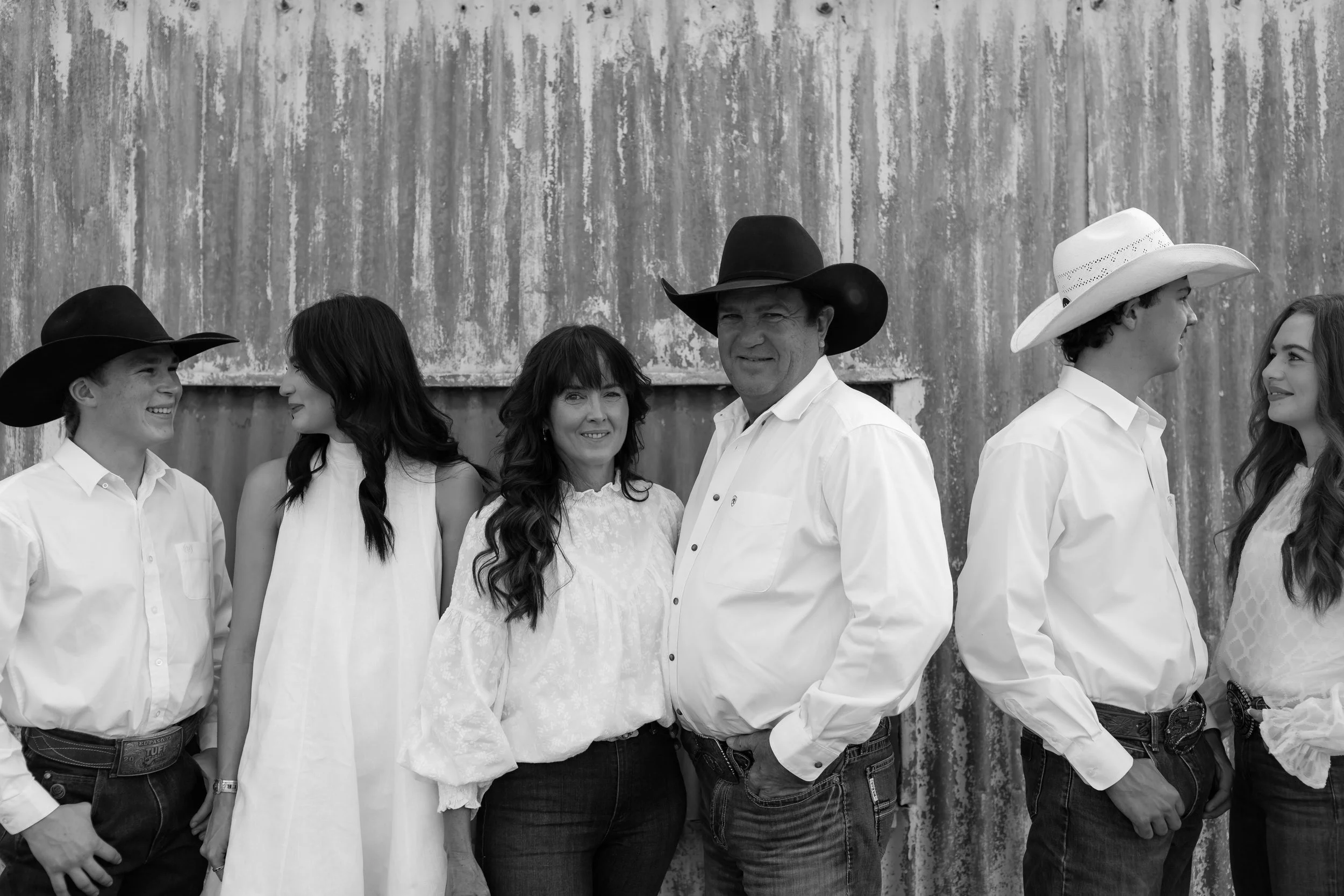 Group of six people dressed in Western attire, standing against a textured metal wall, smiling and interacting with each other.