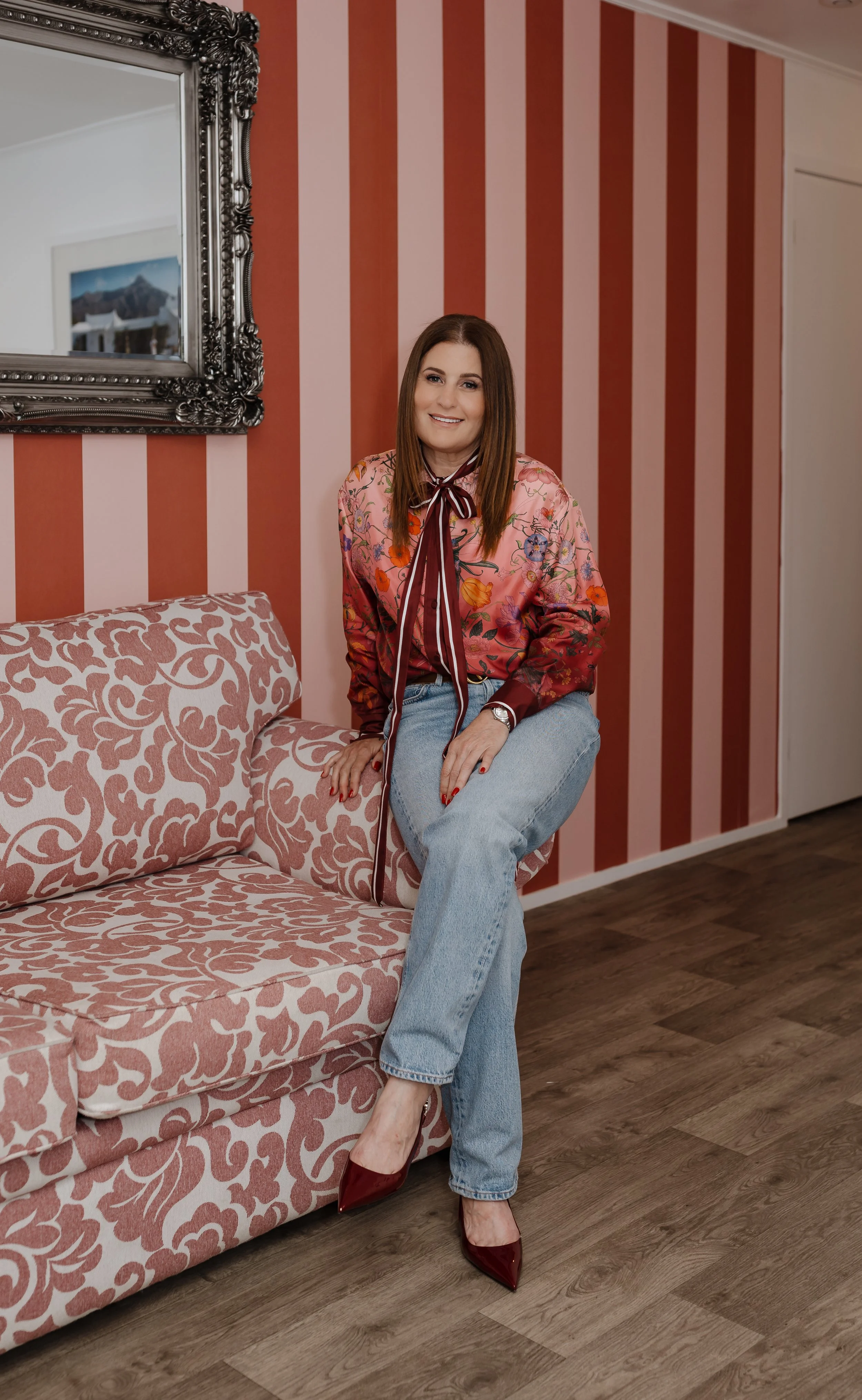 A woman with straight brown hair, wearing a floral silk blouse with a bow collar, light blue jeans, and red high heels, sitting on a pink patterned sofa in a room with pink and red striped wallpaper and a large ornate mirror.