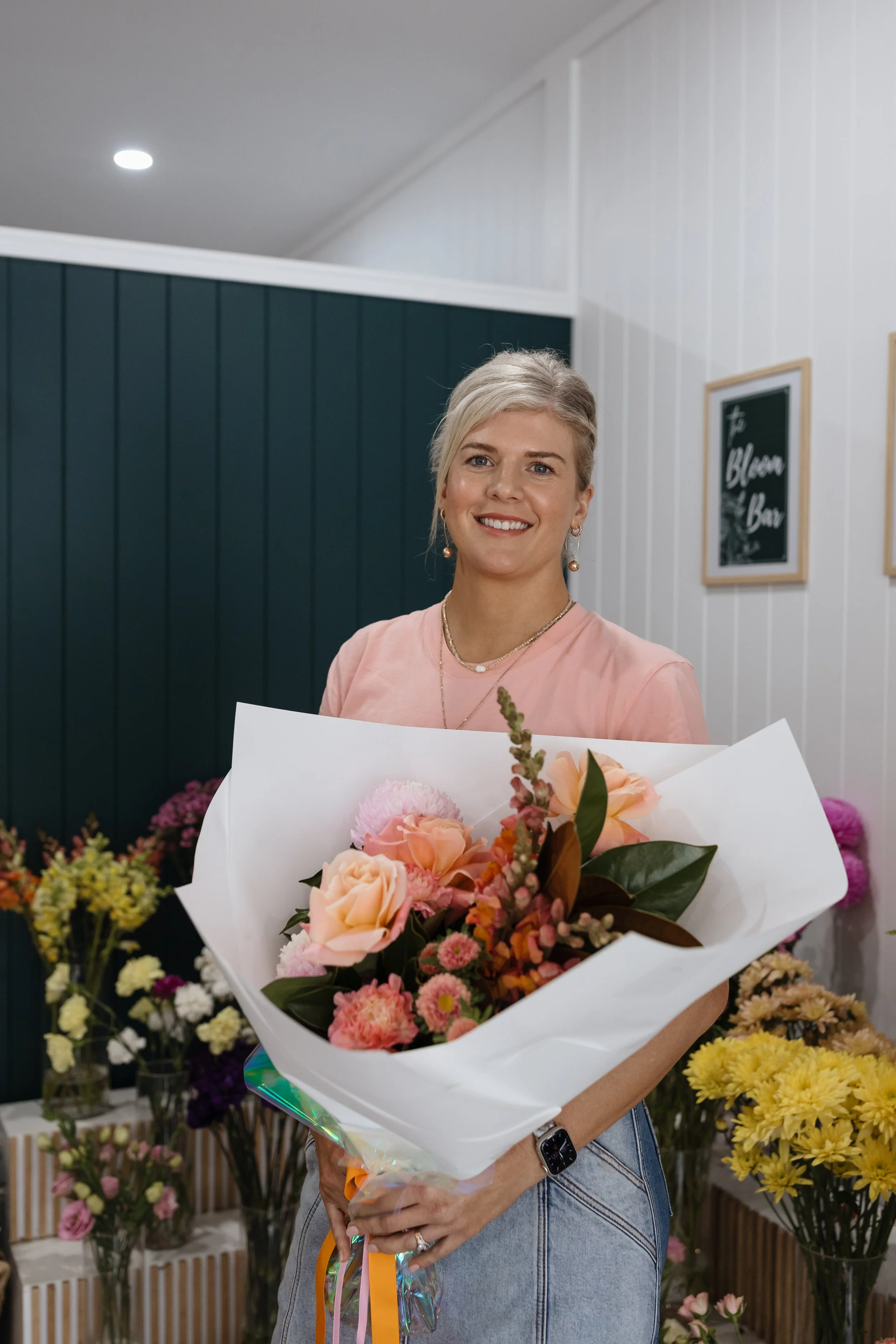 Woman with short blonde hair smiling, holding a bouquet of pink and peach flowers, surrounded by various colorful flowers in a flower shop.