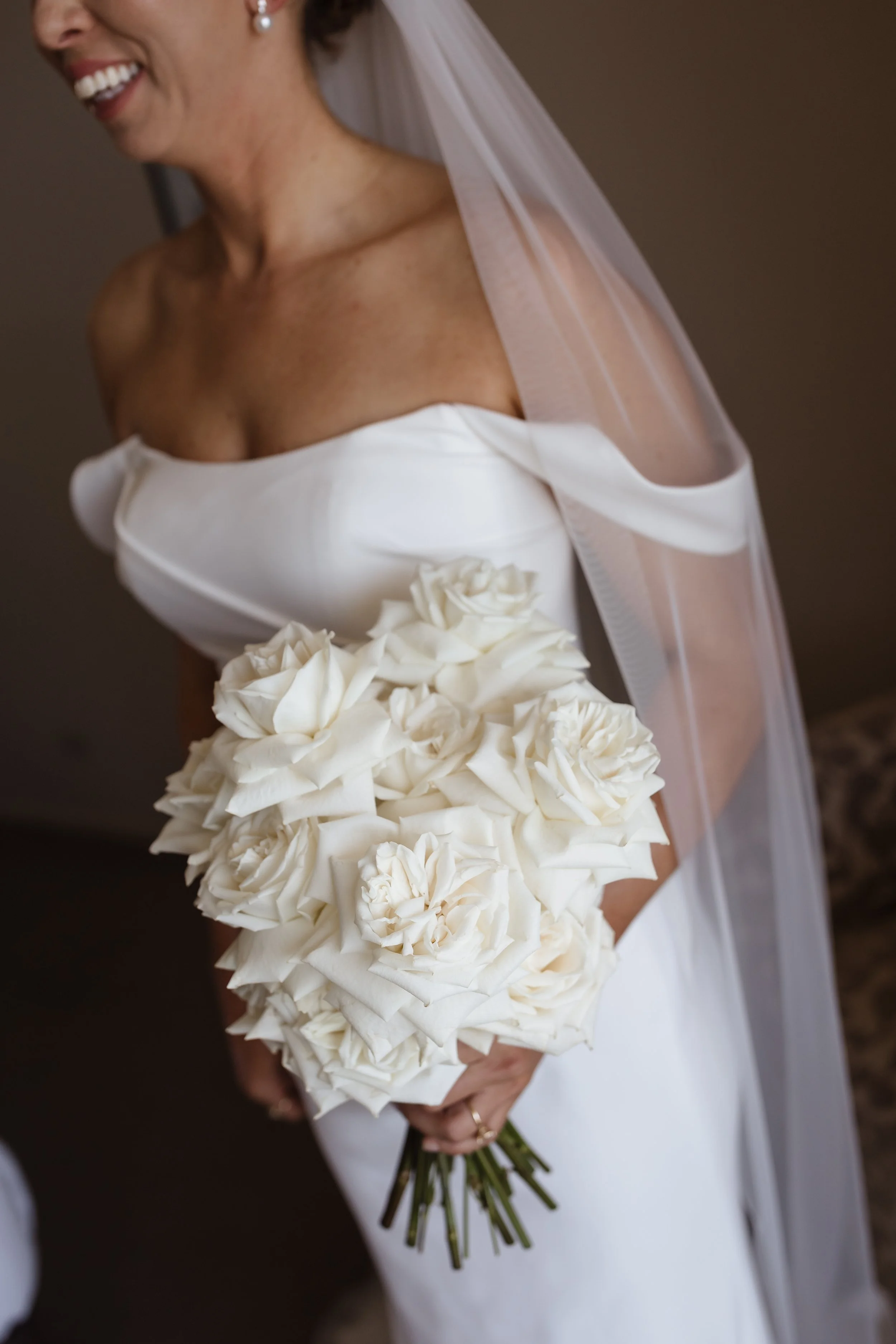 A smiling woman in a white wedding dress holding a bouquet of large white roses.