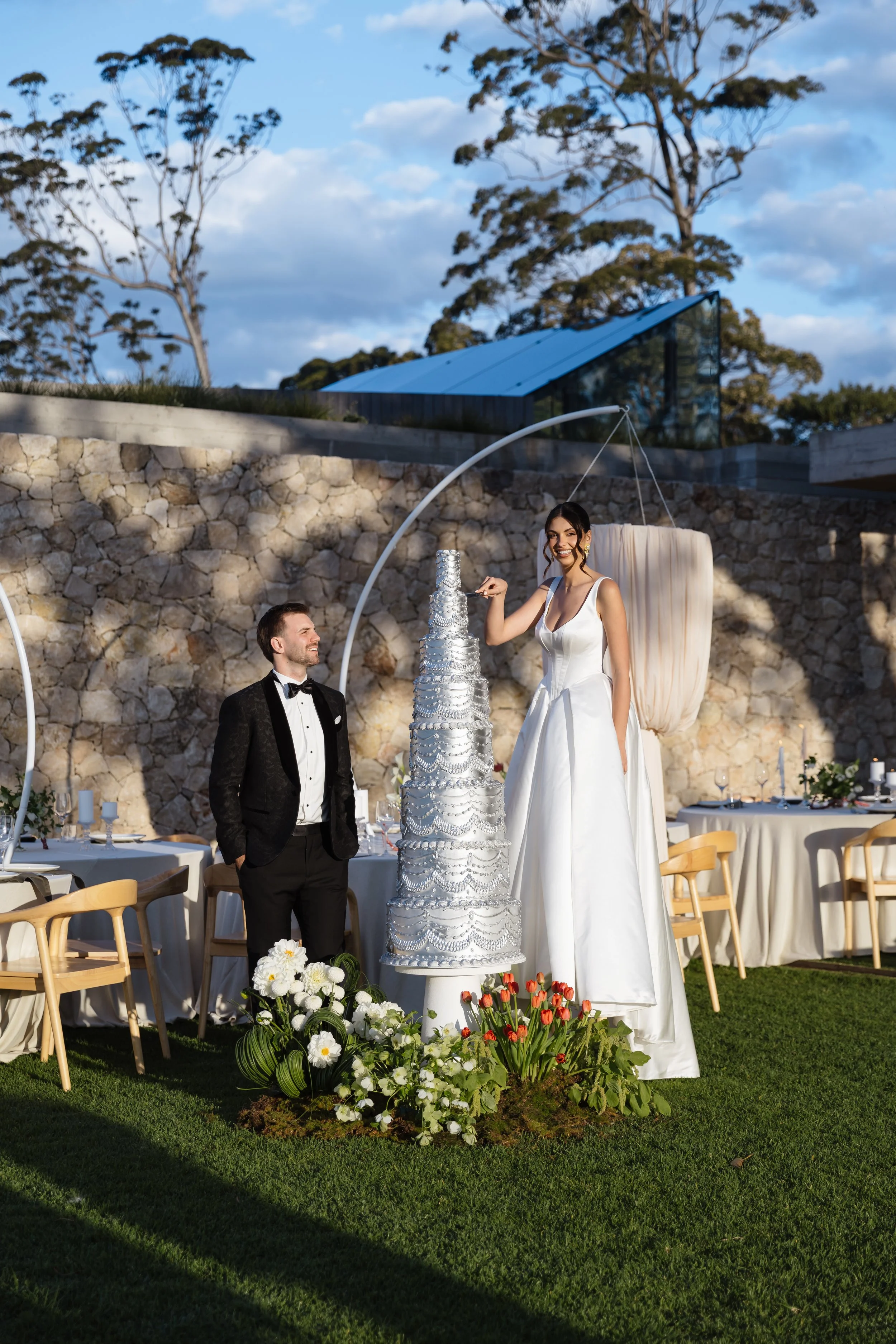 A bride and groom at a wedding celebration outdoors, the bride in a white gown standing next to a multi-tiered wedding cake, and the groom wearing a black tuxedo, with tables, chairs, and floral arrangements nearby, and a stone wall with trees in the