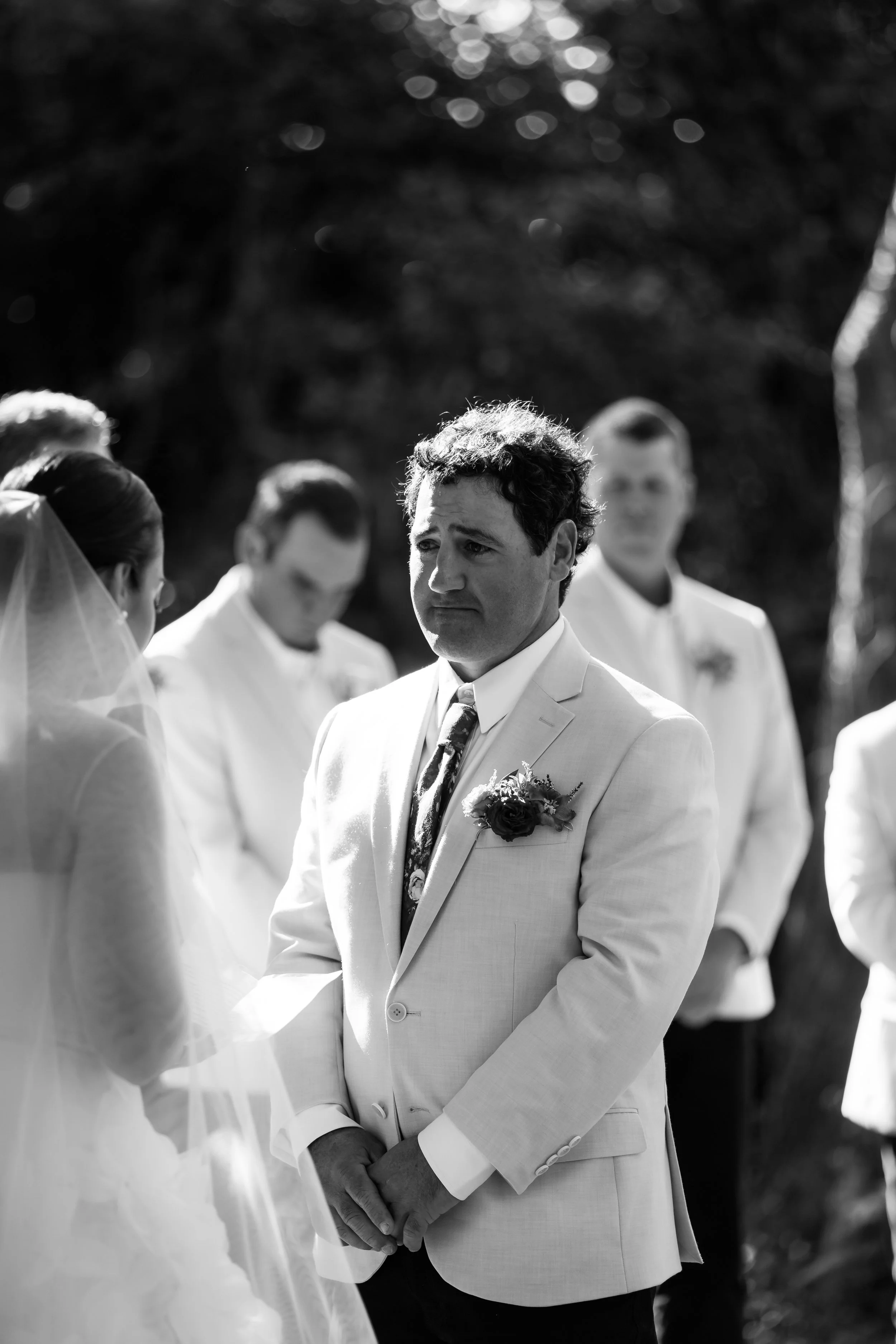 Black and white photo of a groom dressed in a light-colored suit with a floral boutonniere, holding hands with a bride during a wedding ceremony outdoors. Several other men in suits are standing behind him, and a woman in a veil and gown is partially