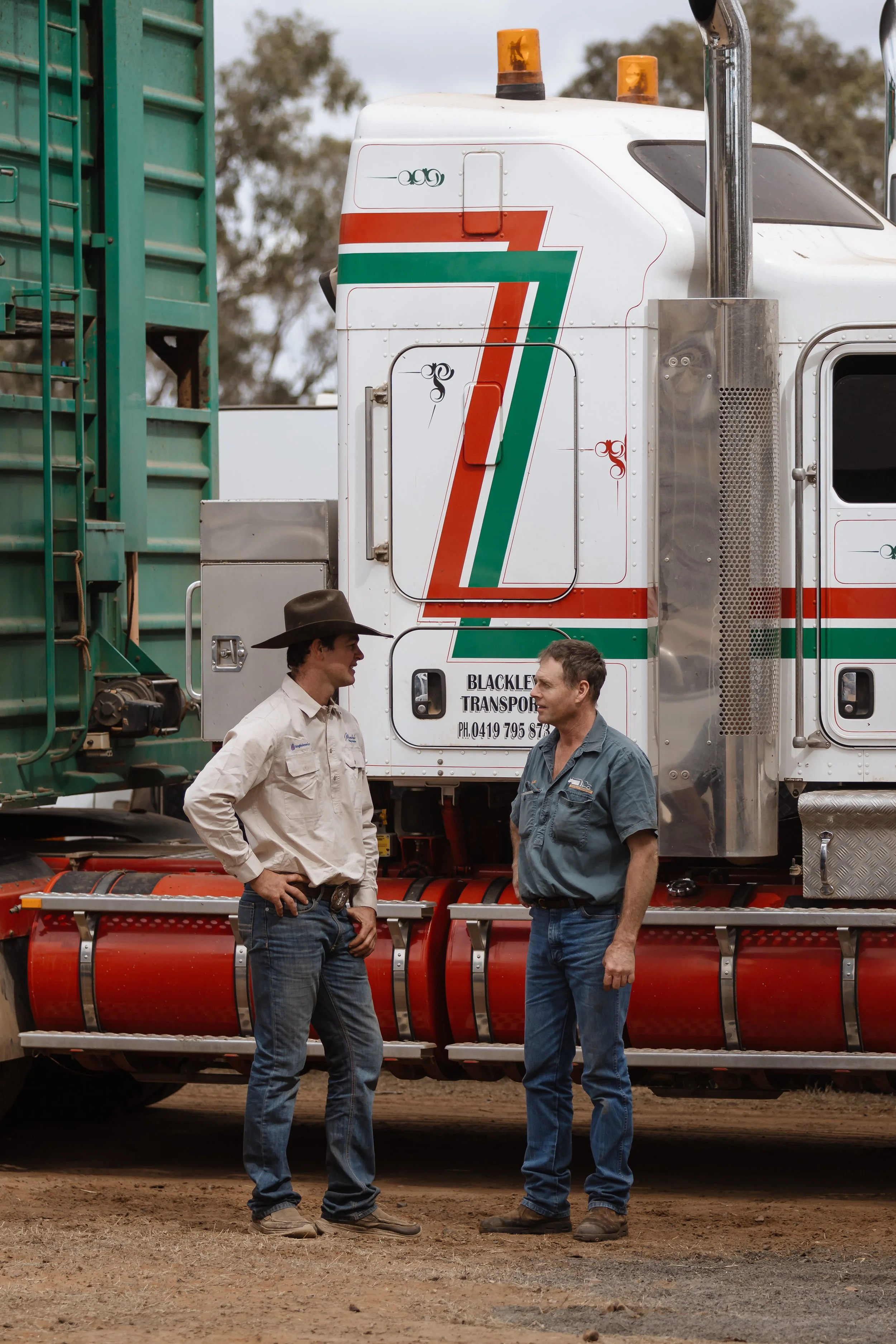 Two men in casual work clothes and hats are standing and talking in front of a large truck. The truck has the number 7 painted on it with red, white, and green stripes, and there are orange warning lights on top. The background shows blurred trees an
