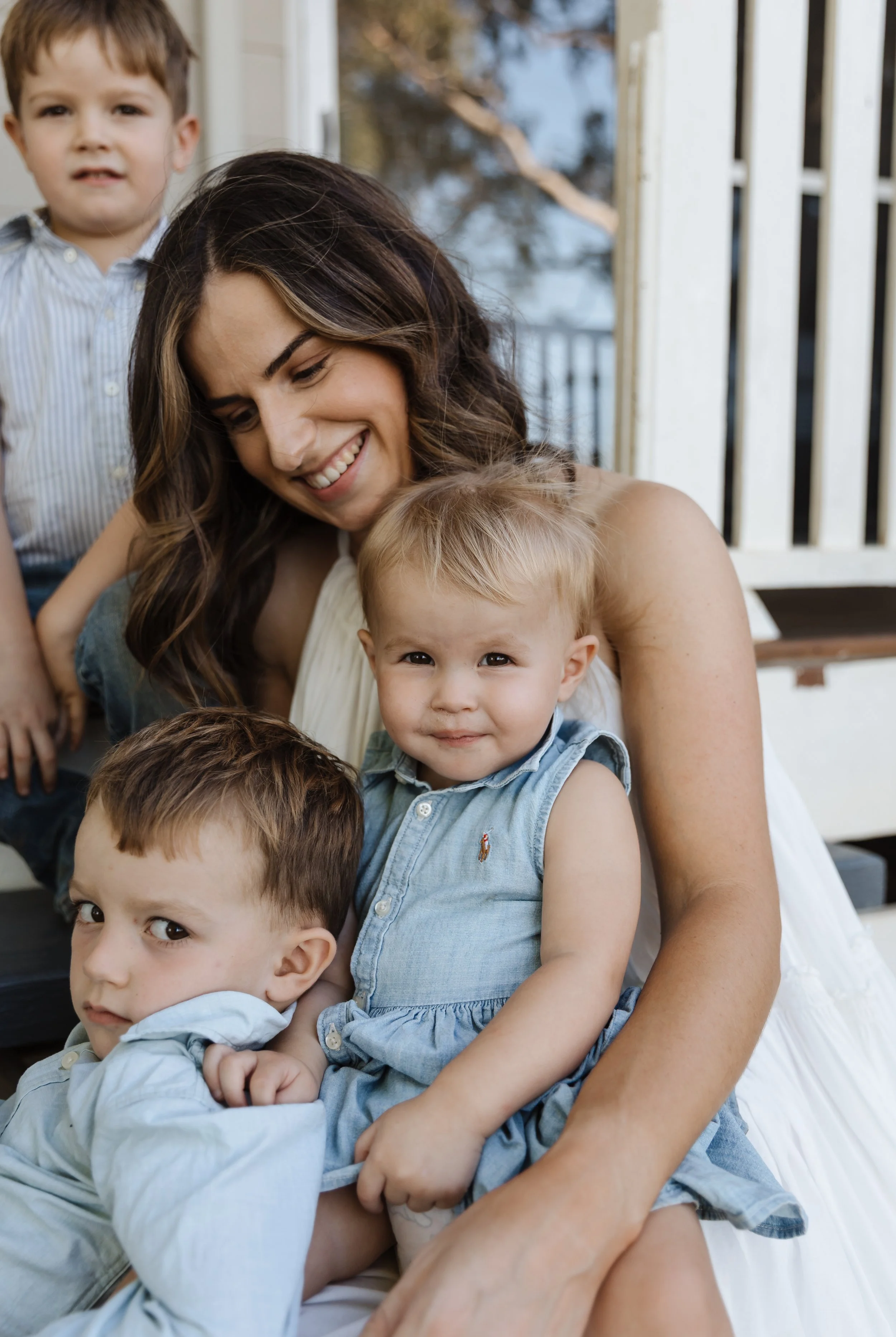 A woman with long dark hair and a white dress smiling while cuddling four young children with light hair and blue or gray shirts on a porch.
