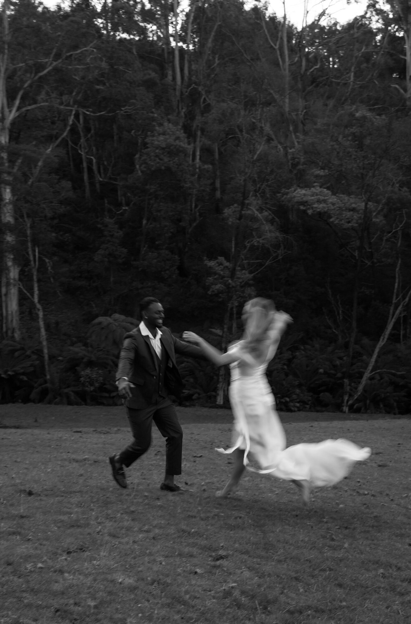 A couple dancing outdoors in a wooded area, holding hands and spinning together. The man is wearing a suit, and the woman is wearing a long dress. The image is in black and white.