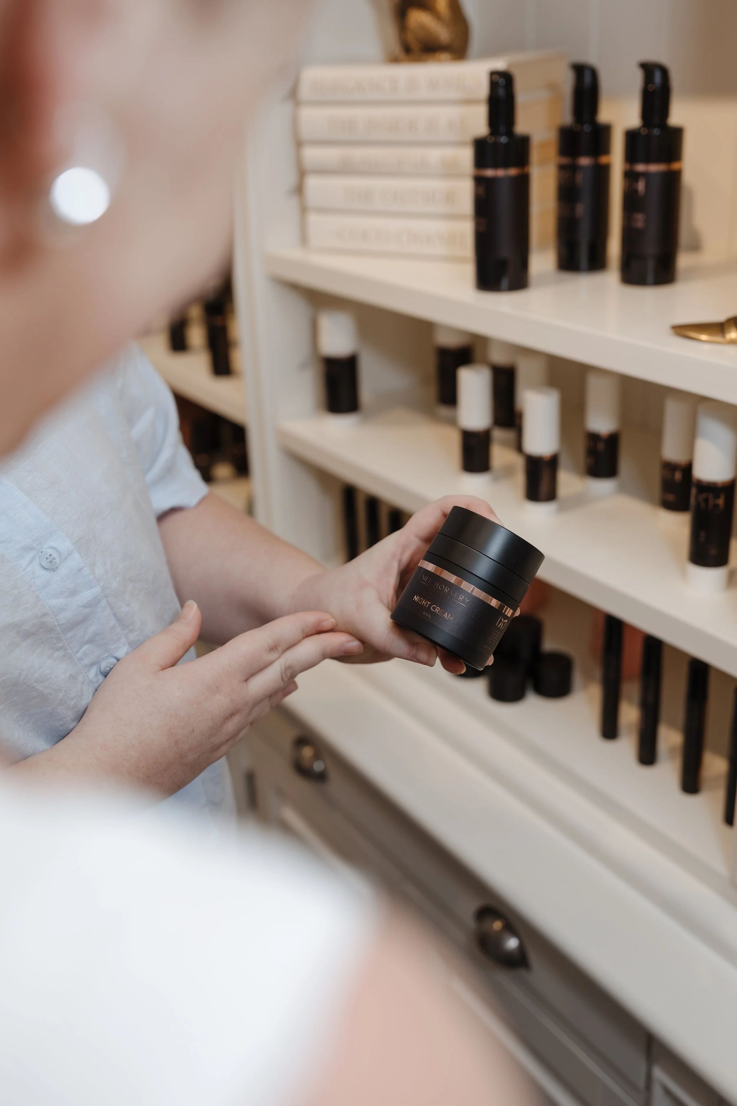 A woman is holding a black container labeled 'Night Cream' while standing in front of shelves filled with skincare products in black and white bottles.