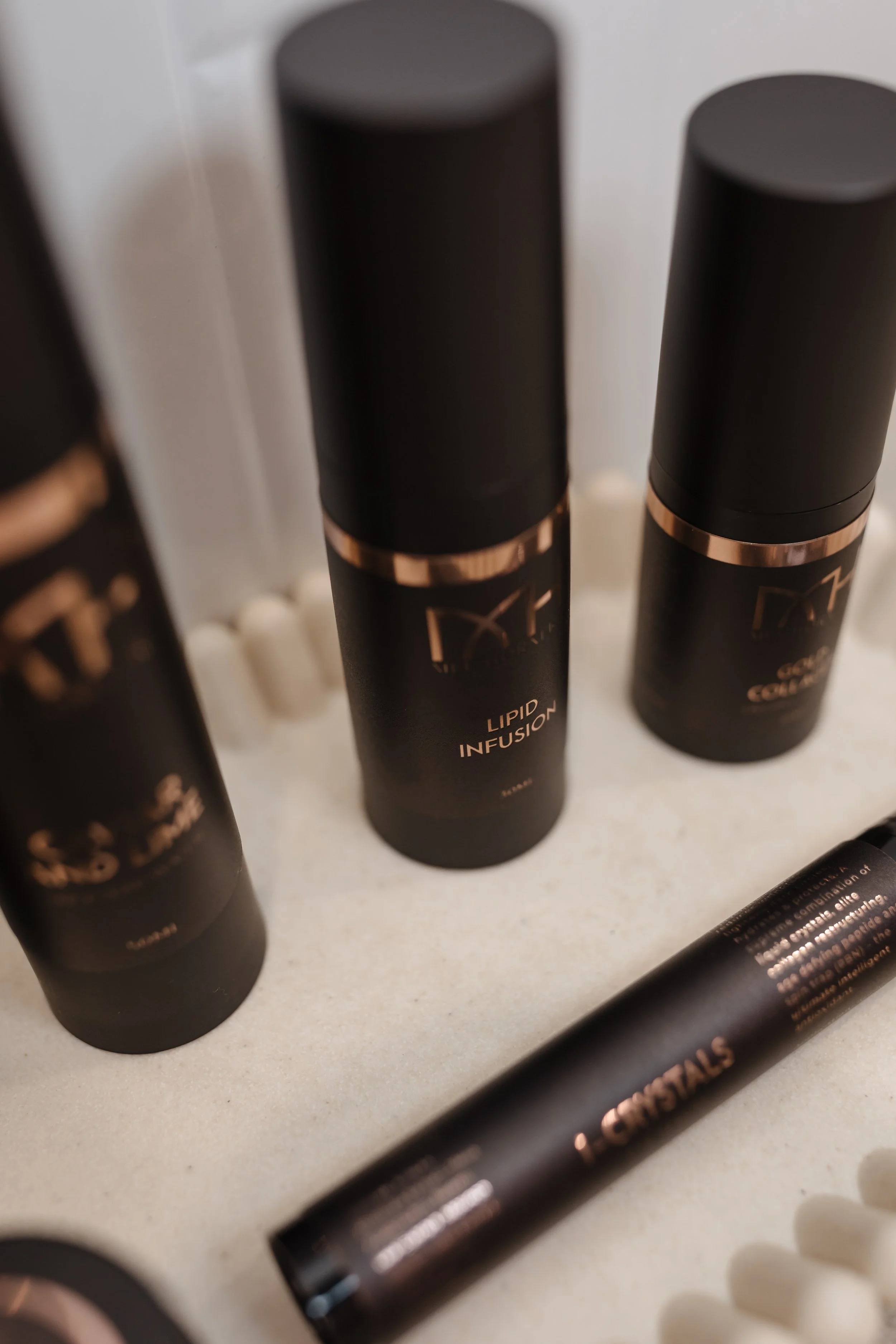 Several black bottles with copper accents labeled 'Lipid Infusion' are arranged on a light-colored surface, with some partial view of other cosmetic or skincare products.