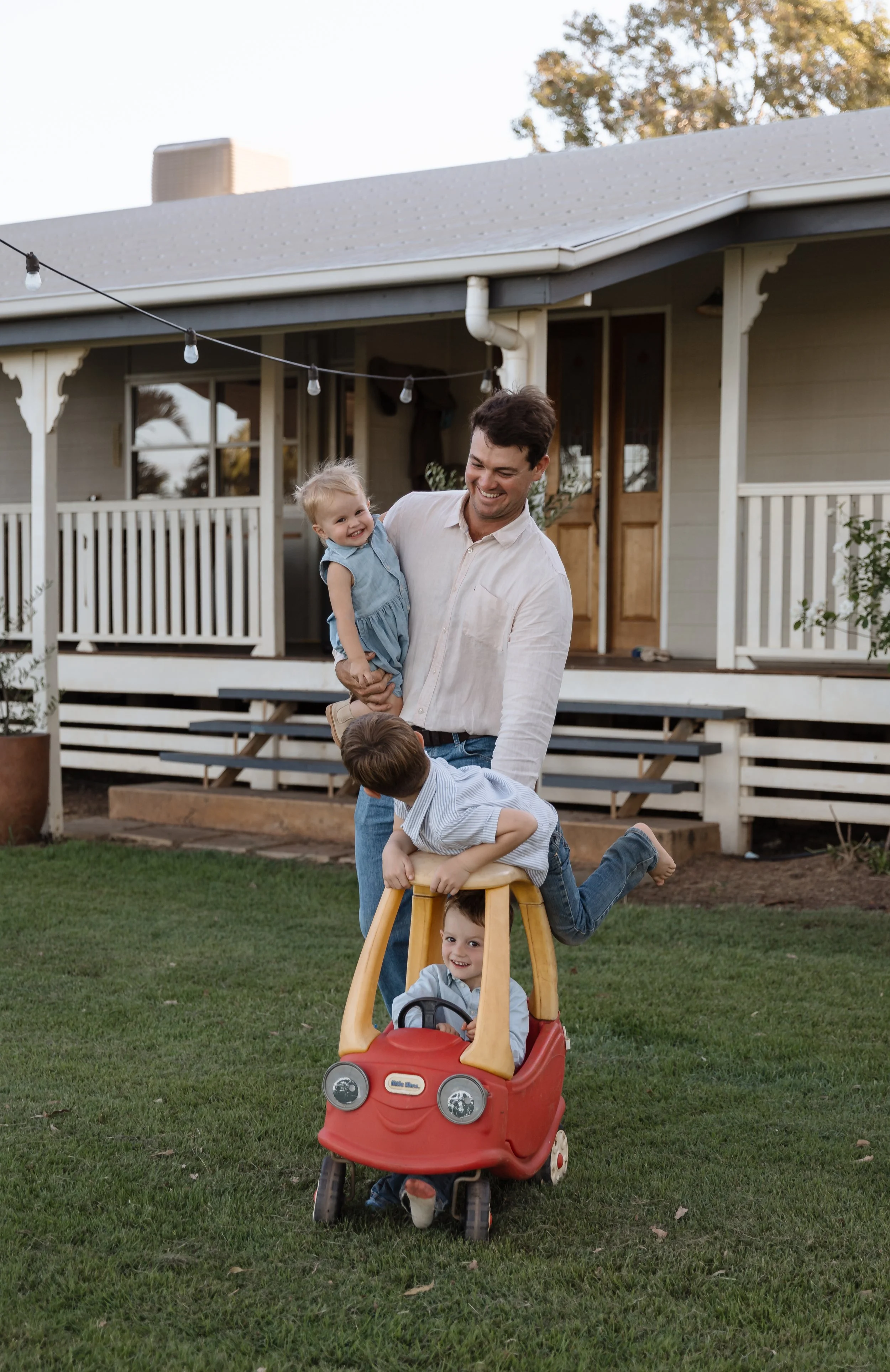A father playing with three children outdoors in a backyard, with a house in the background. One child is sitting in a red toy car, another is riding on a wooden toy with the father, and a girl is being held up by the father and smiling.