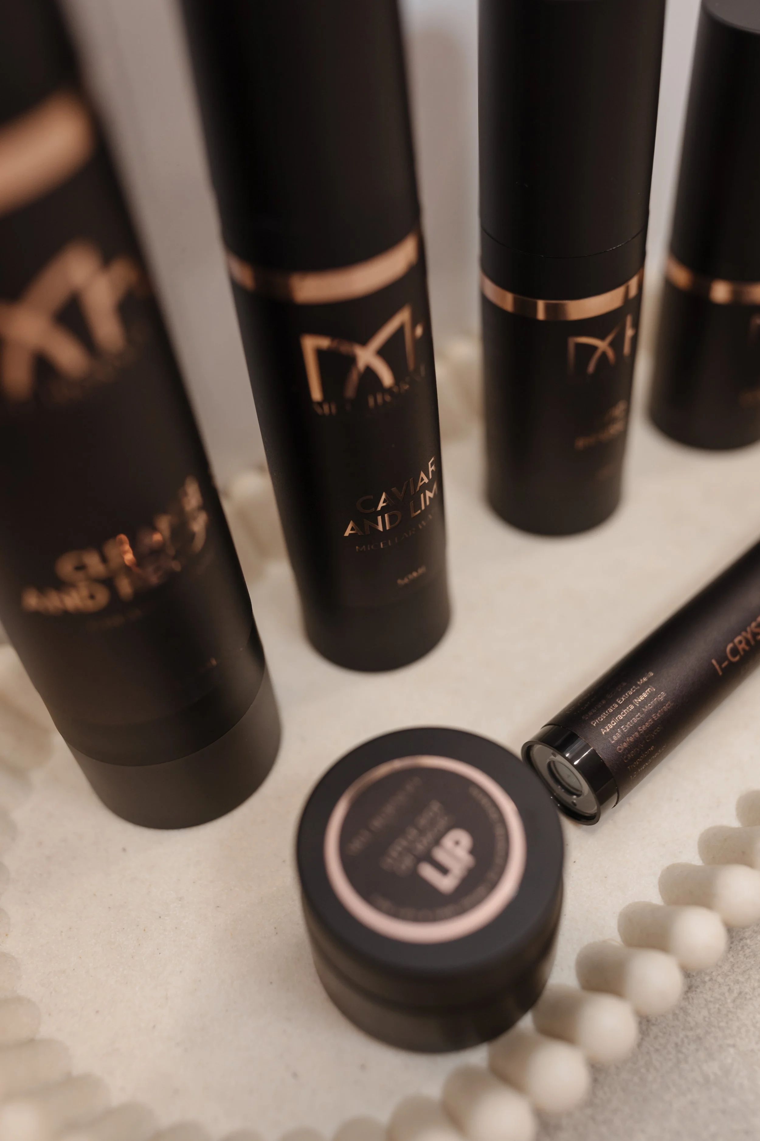 Close-up of black beauty products with rose gold lettering and accents, including tubes and jars, arranged on a light-colored surface with cotton swabs nearby.