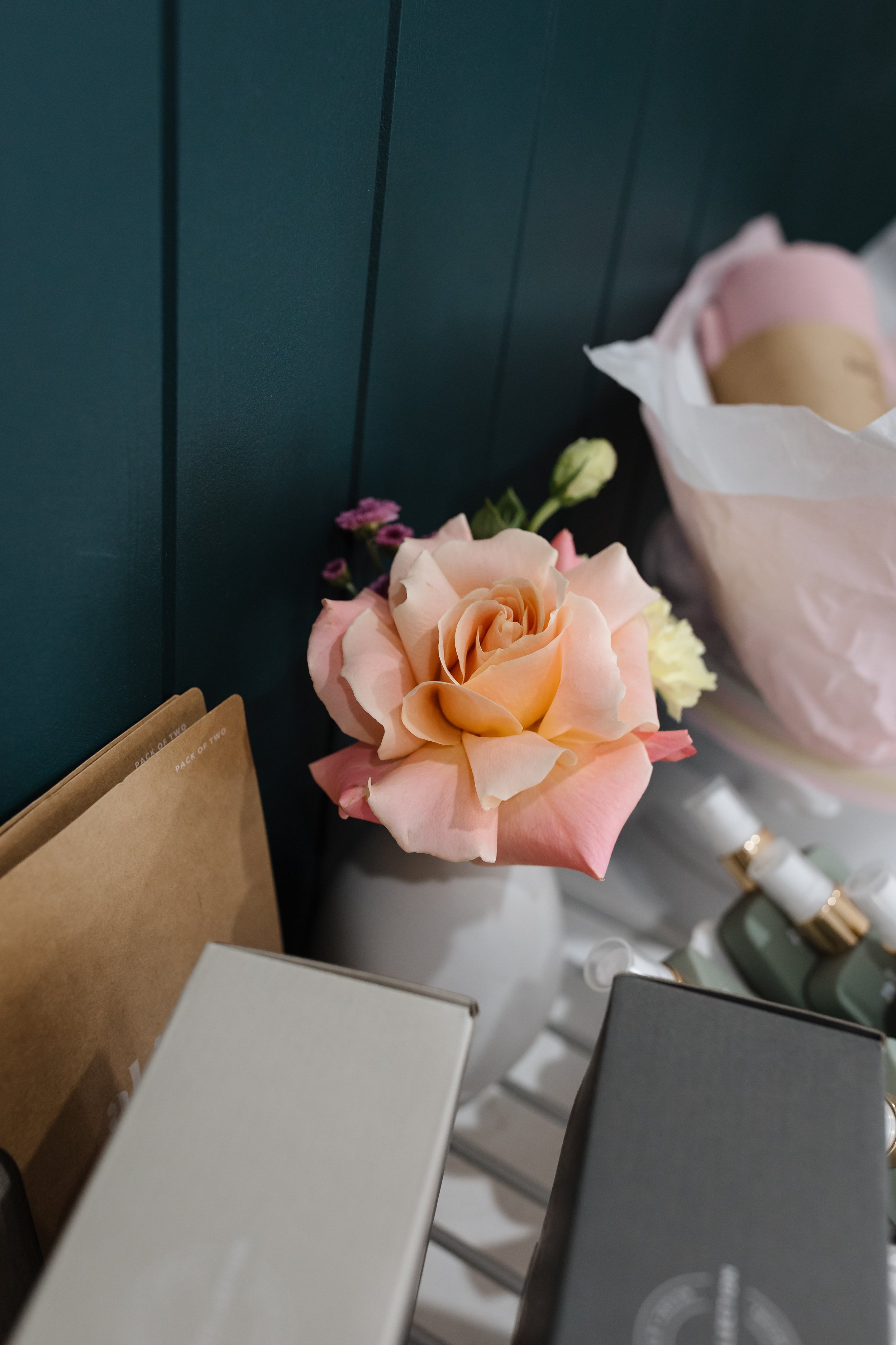 A pink and peach-colored rose with some small purple and white flowers nearby, placed on a surface next to a dark green wall with vertical paneling.