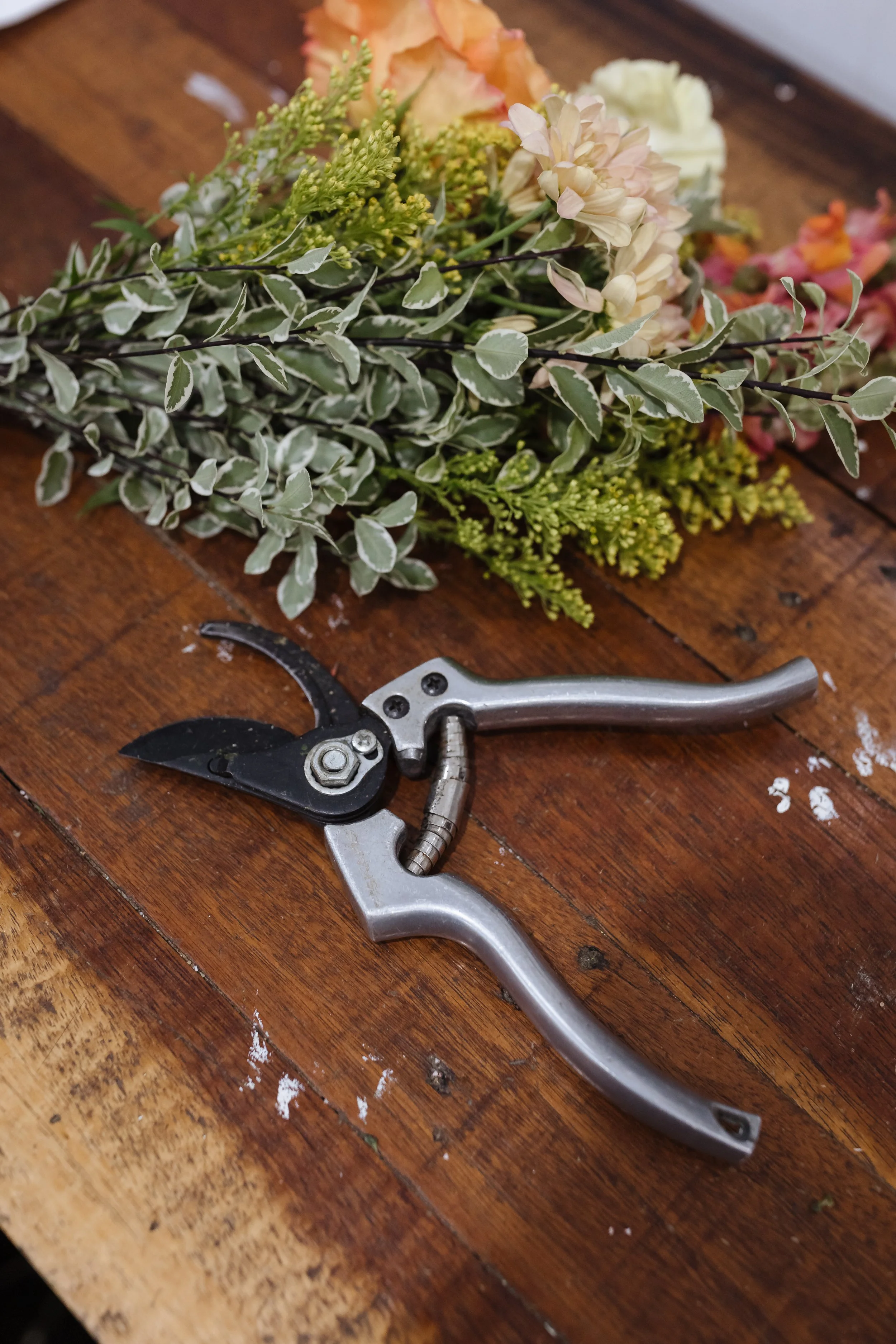 A bouquet of various flowers and greenery on a wooden surface with pruning shears nearby.