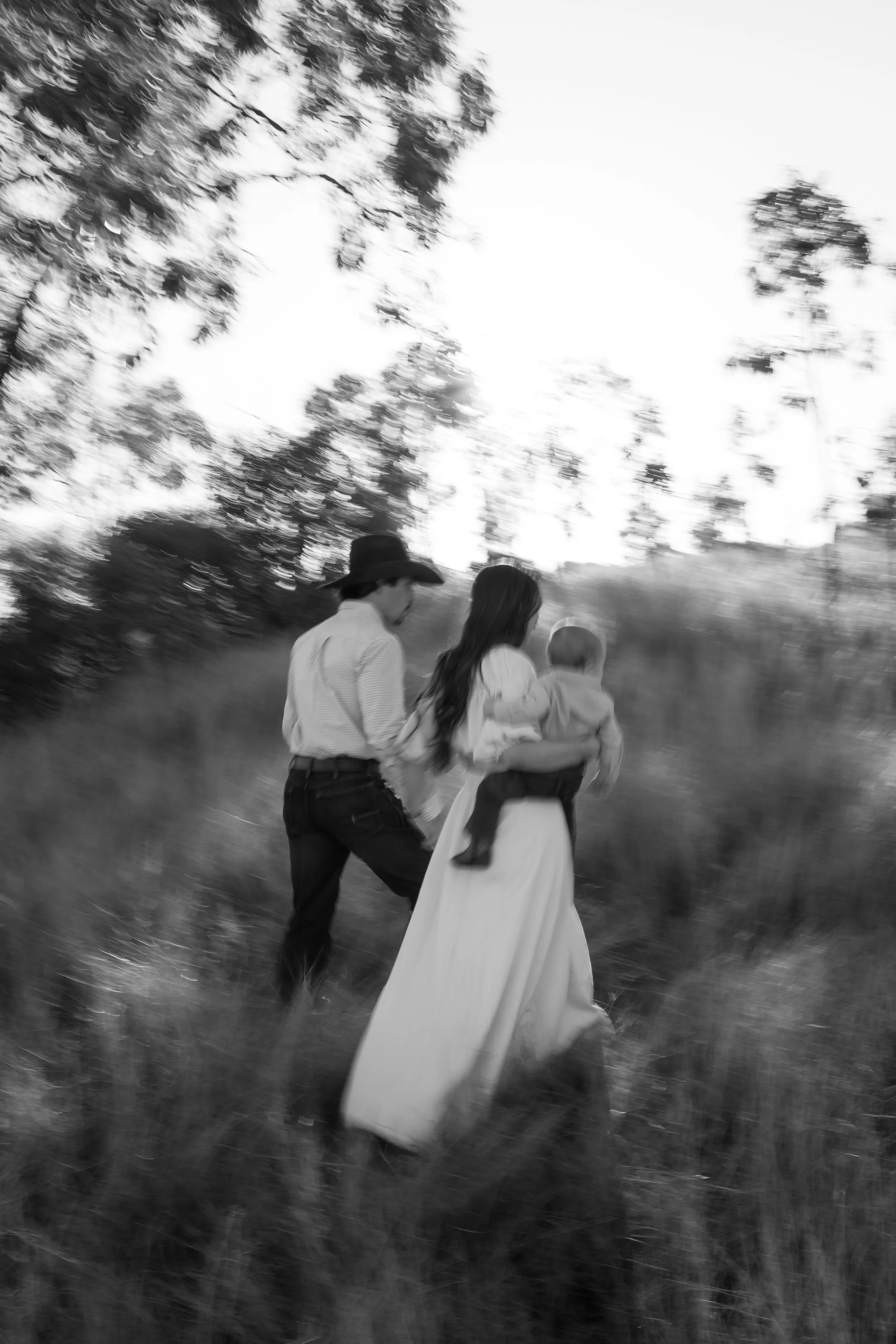 Black and white photo of three people walking outdoors. The person in the center is a woman with long hair, wearing a wedding dress. The person on the left is a man with a hat, and the third person is a young child. The background shows trees and a b