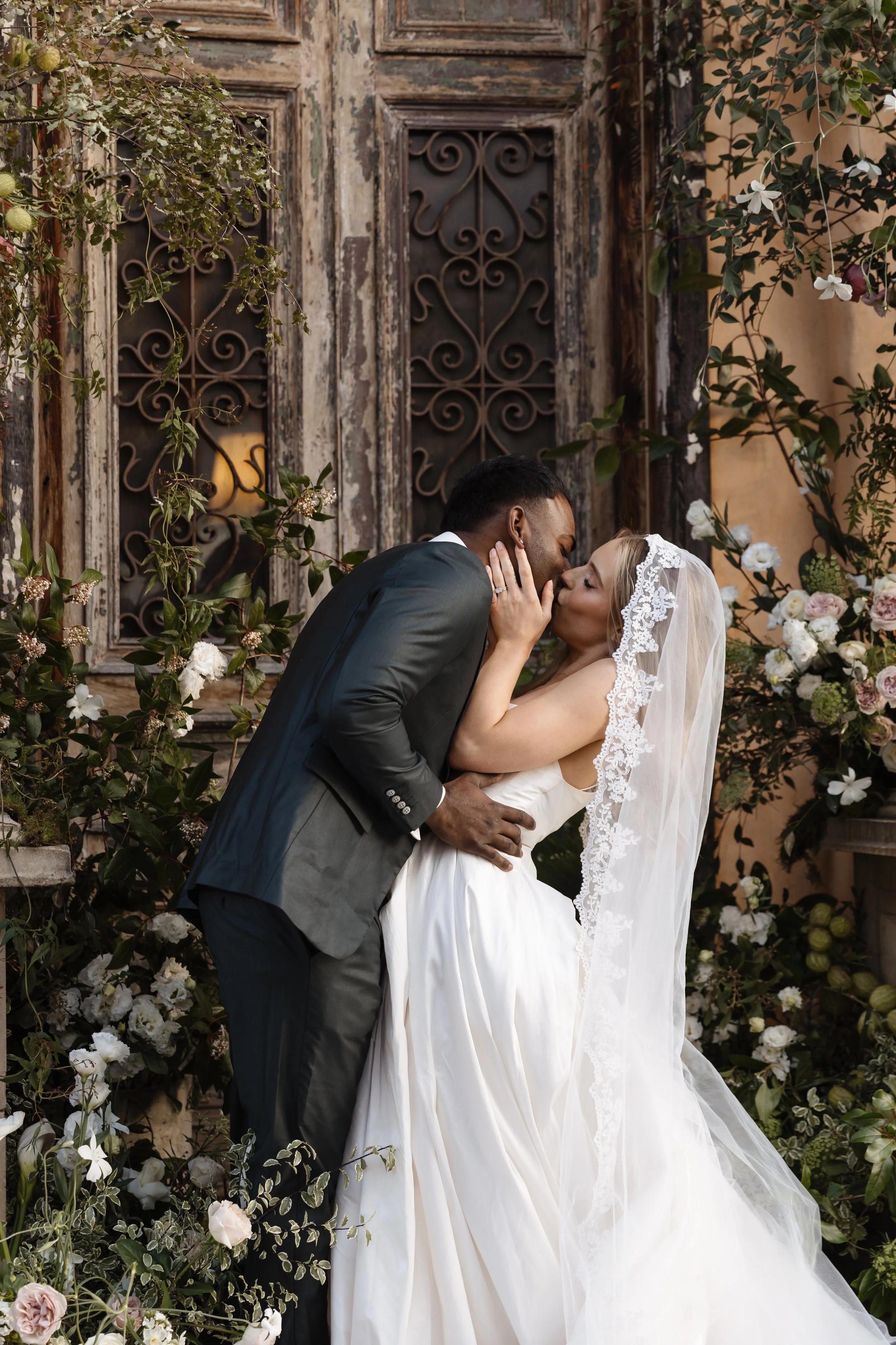 A bride and groom sharing a kiss at their wedding surrounded by flowers and greenery.