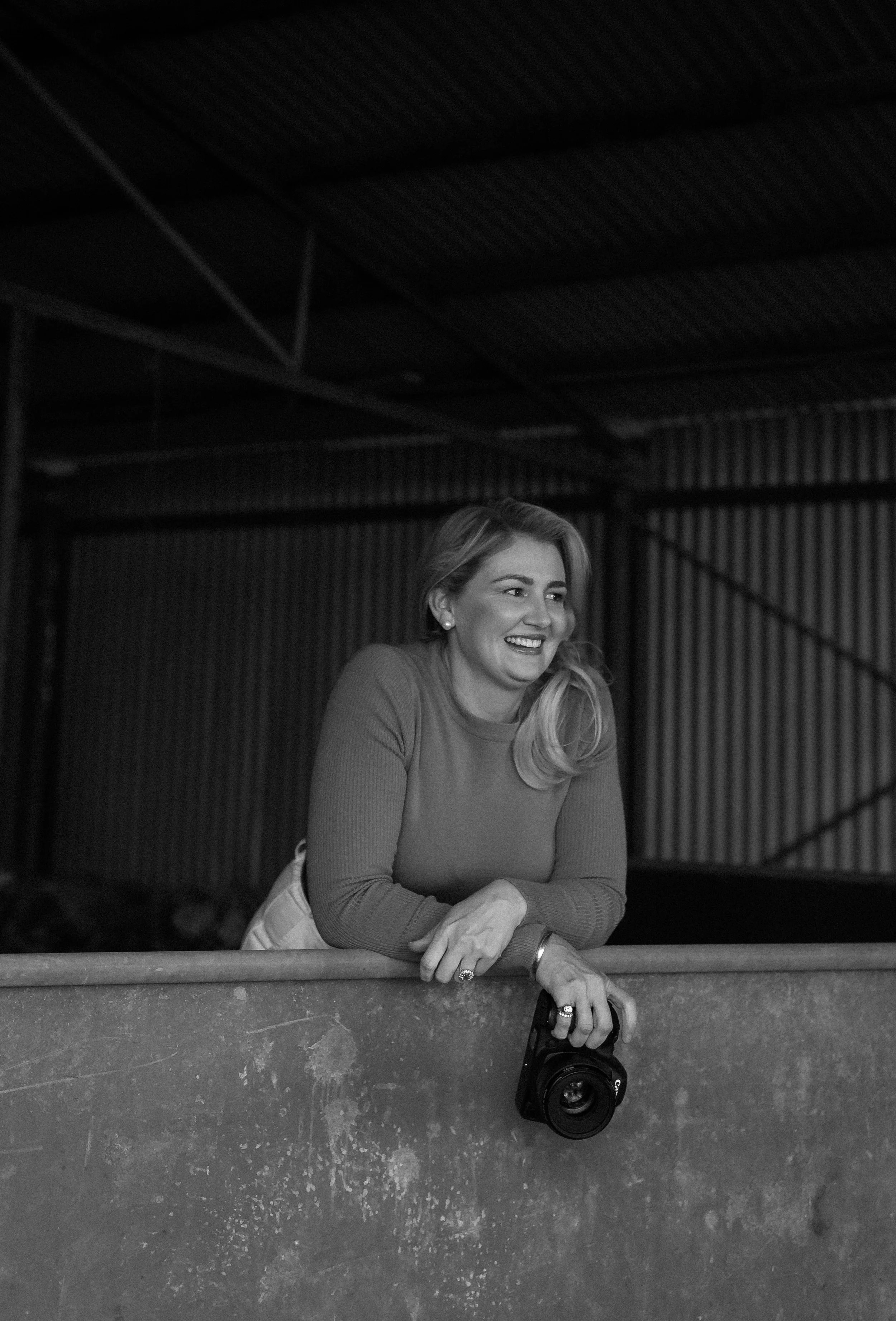 A woman with long wavy hair smiling, leaning on a ledge with a camera in front of her on the ground. She is wearing a long-sleeved shirt and has jewelry on her wrist and finger. The background appears to be an industrial or warehouse-like setting with a metal and wood ceiling.