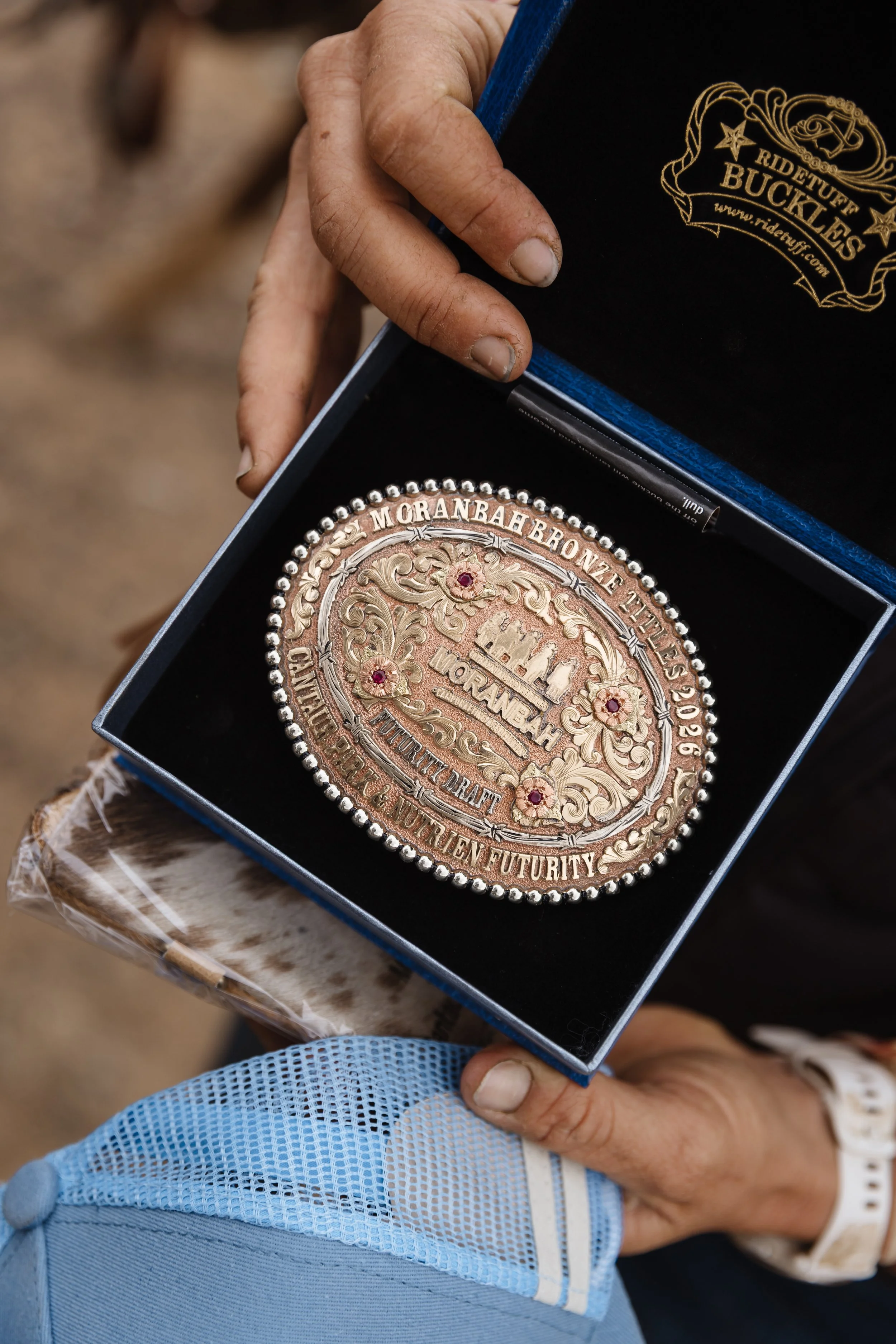 Close-up of a person's hand holding a decorative medal with intricate designs and inscription, inside an open box with a logo for Ride Tuff Buckles. The person is wearing a black shirt with a gold logo, and another hand with a blue cloth is visible.