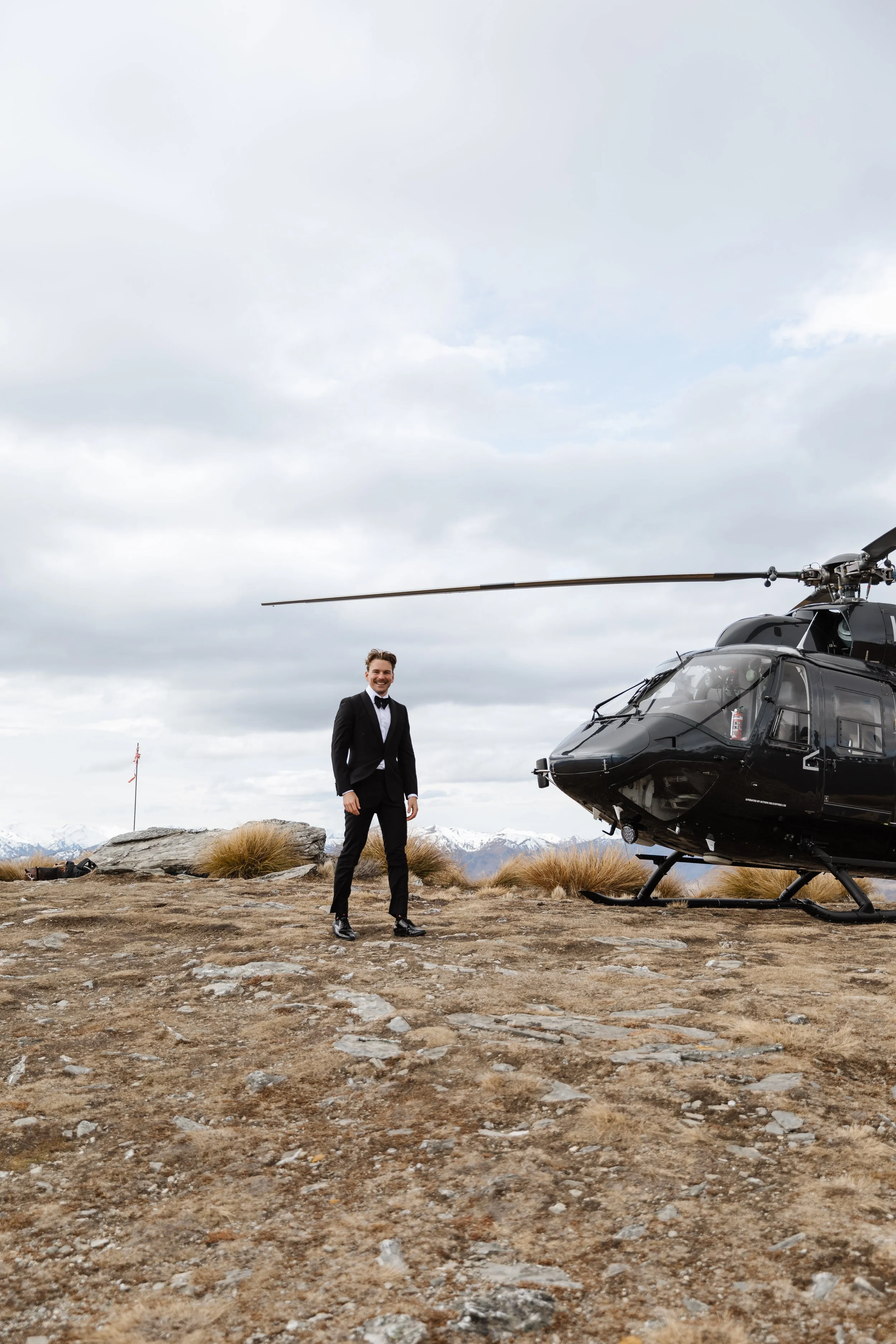 A man in a tuxedo standing outdoors on rocky terrain in front of a black helicopter with snow-capped mountains in the background.