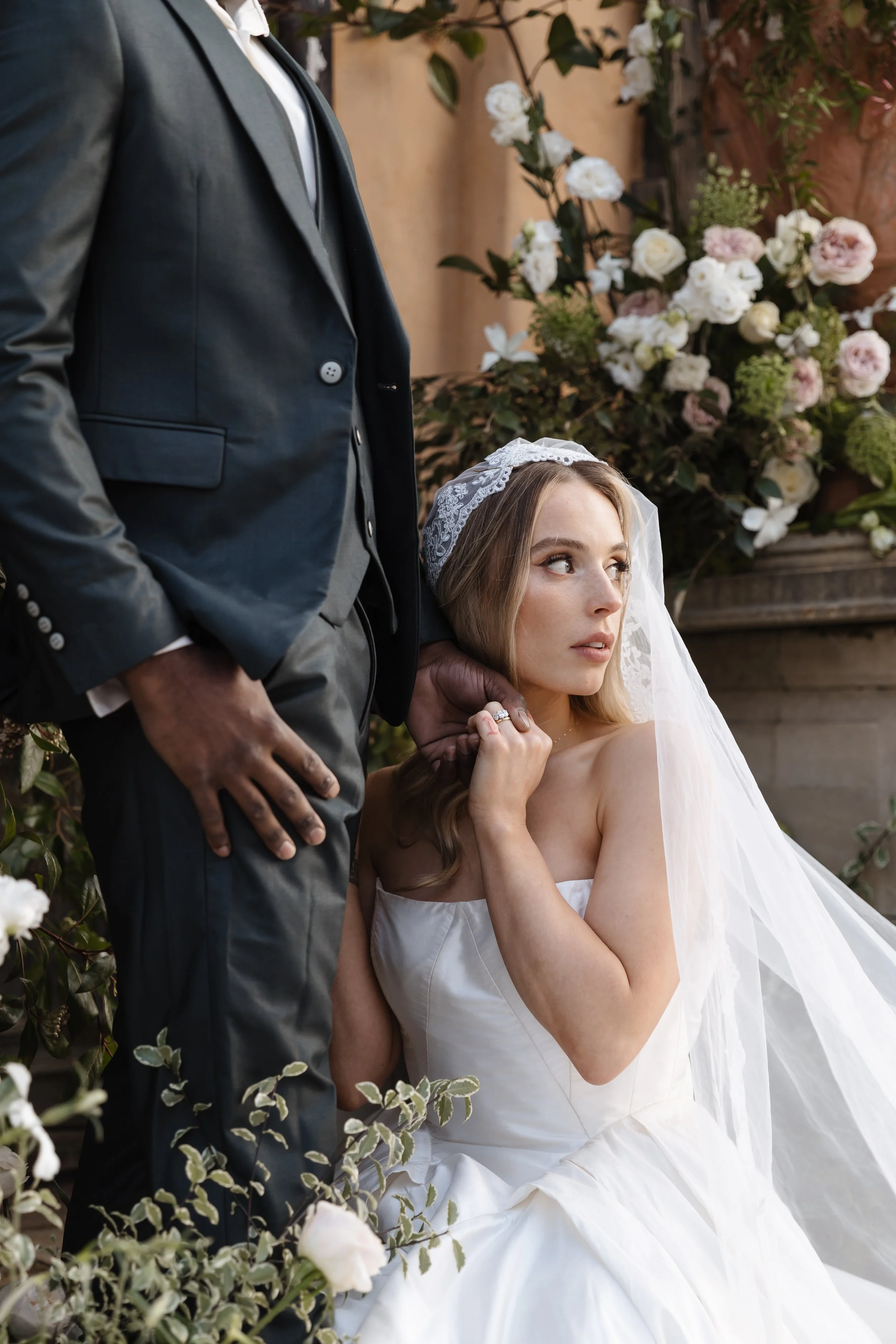 A woman wearing a white wedding dress and veil is sitting, holding her chin with her hand, while a man in a black suit stands beside her. They are surrounded by a lush floral arrangement with white and pink flowers and green foliage.