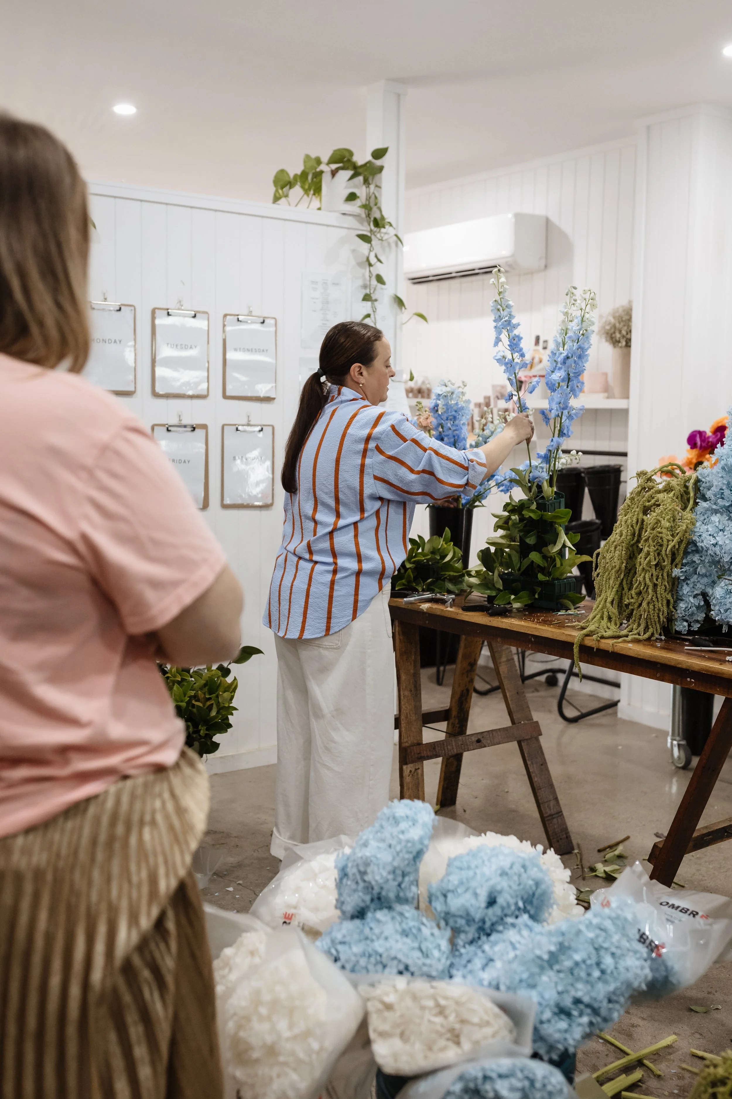 A florist arranging blue and white flowers in black vases in a flower shop, with bundles of flowers and floral foam on the floor.