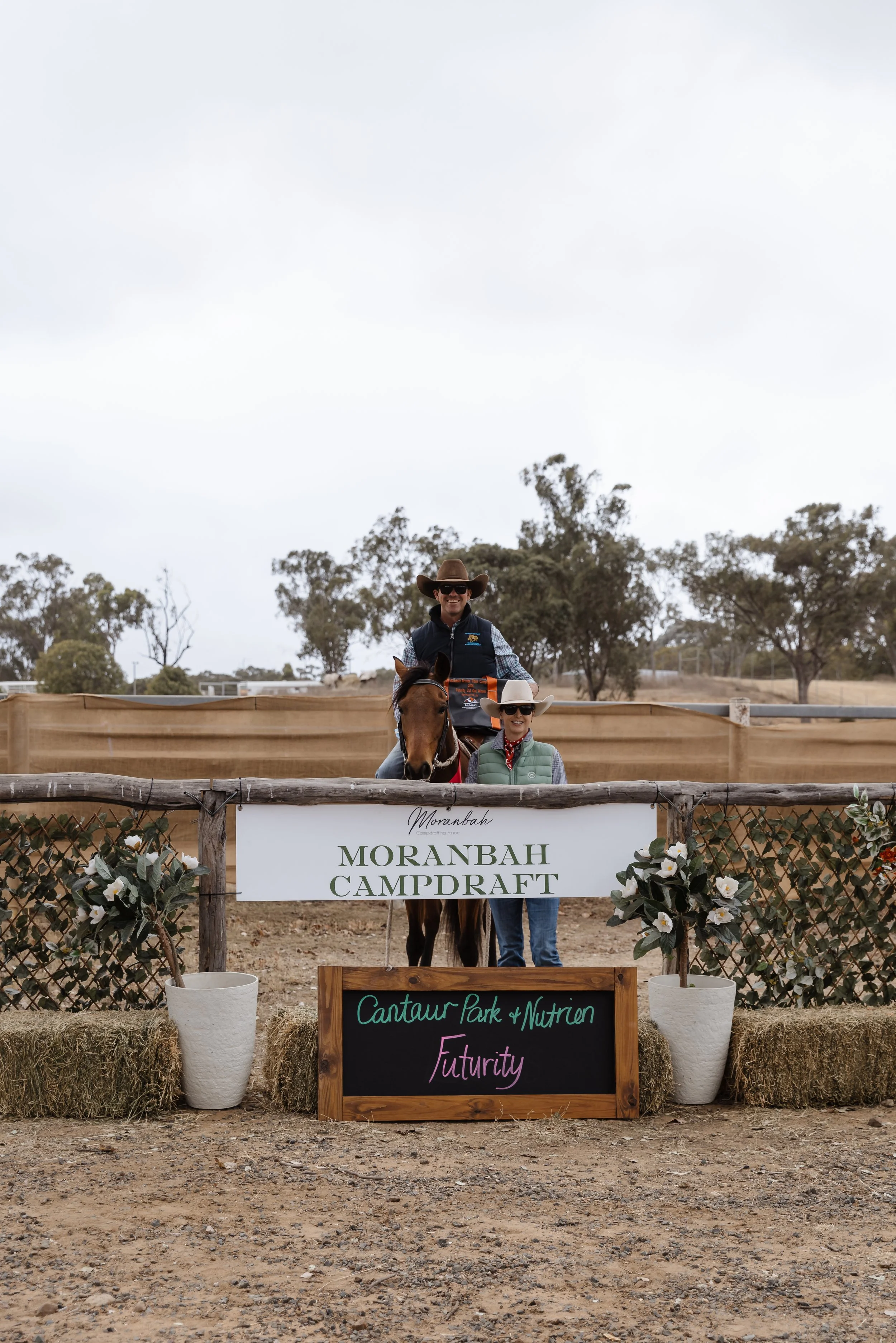 Two people on a horse at an outdoor event with a banner reading 'Moranbah Campdraft'. There are trees in the background and decorative flowers around the sign.