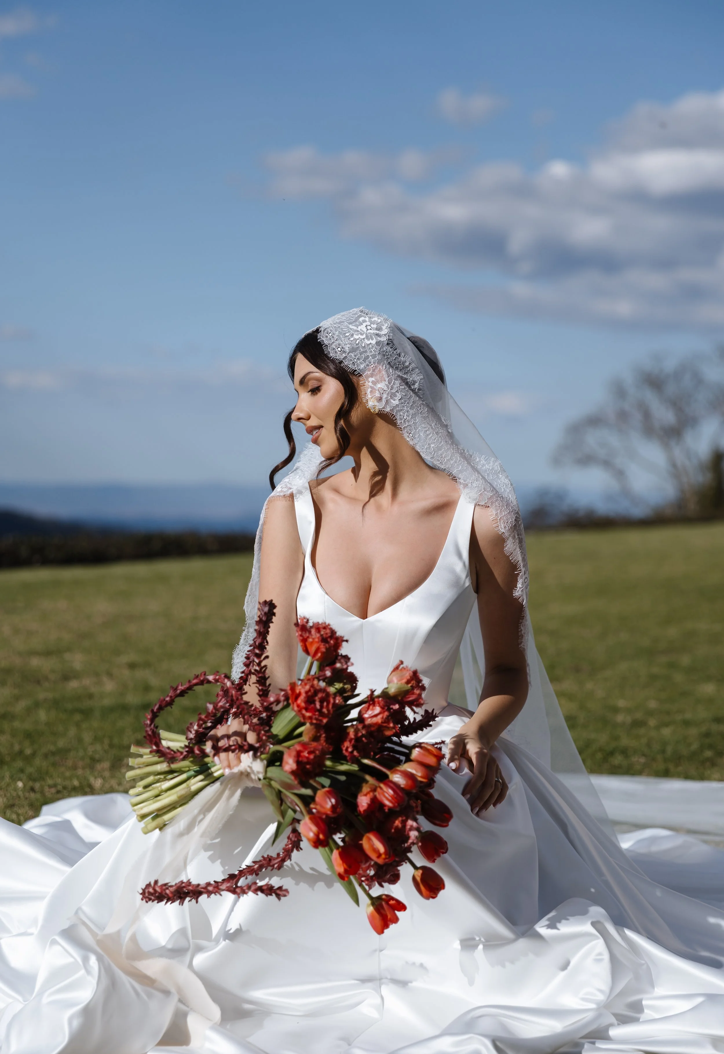 A bride in a white wedding dress with a lace veil sitting outdoors on grass, holding a bouquet of red and orange flowers, with a blue sky and scattered clouds in the background.