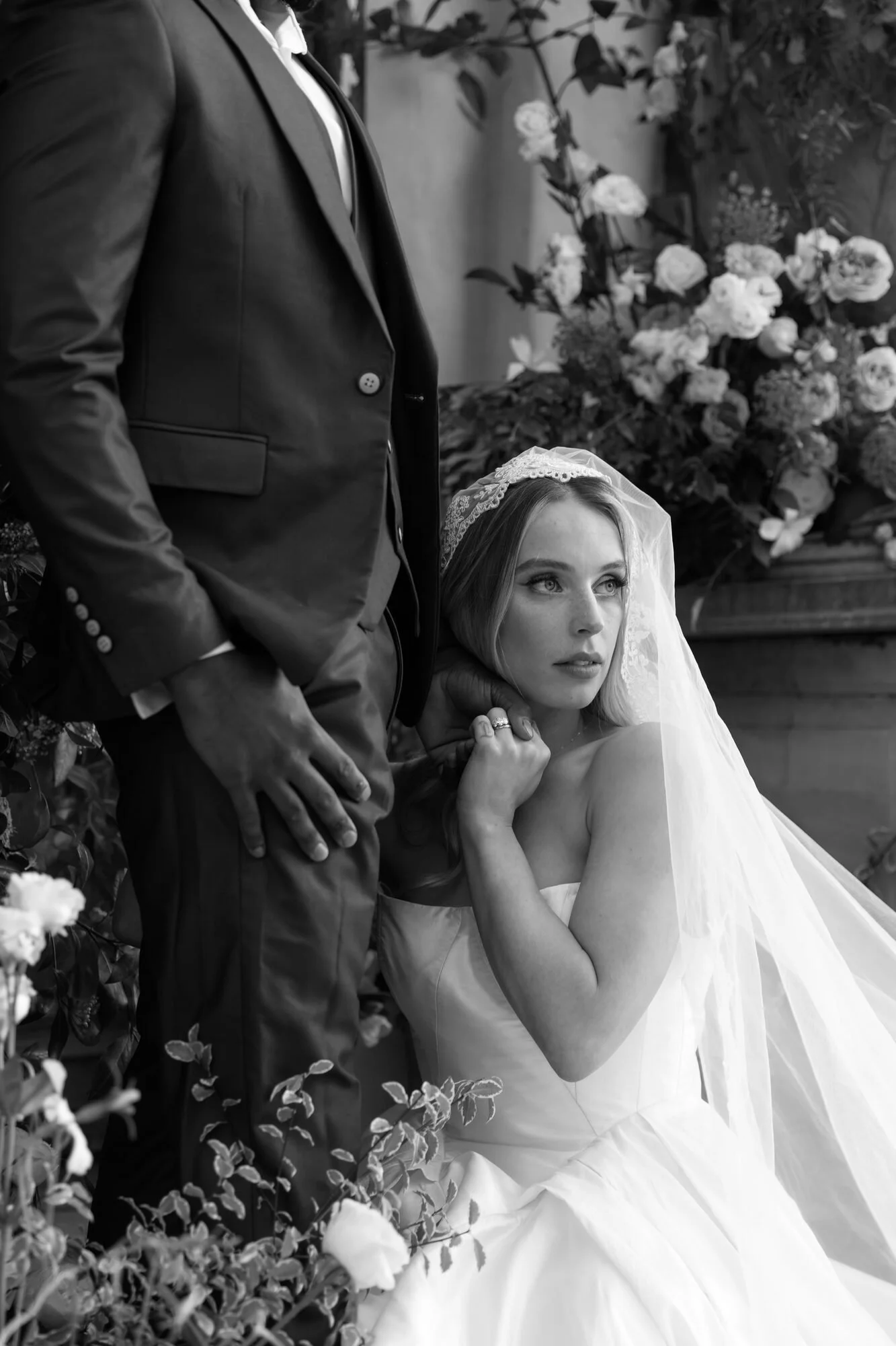 A bride with a lace veil and strapless gown resting her head and hand on a groom's chest, who is wearing a suit jacket, with a background of flowers and greenery.