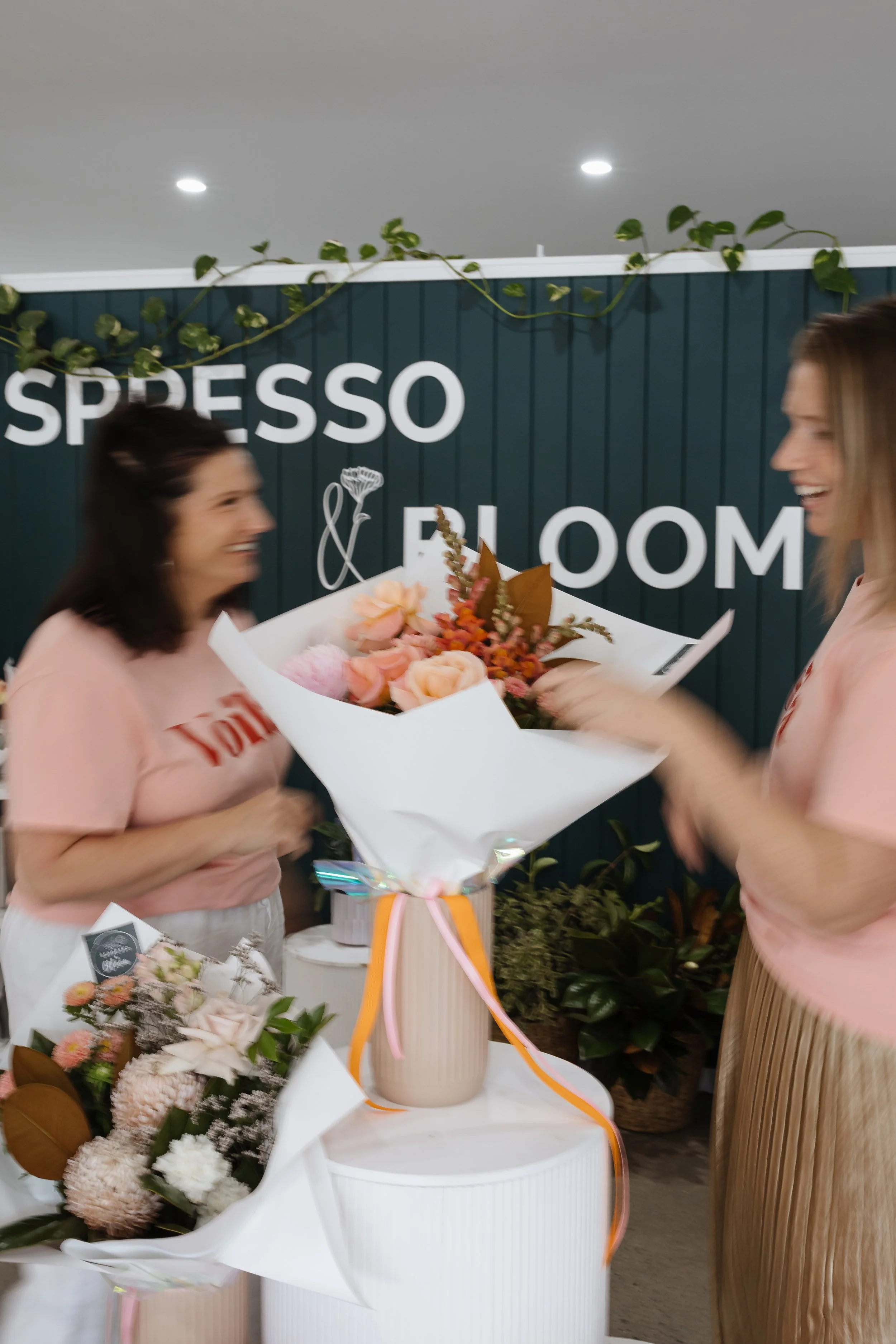 Two women smiling and exchanging a bouquet of flowers in a shop or market with a dark green wall and white sign that says 'Espresso & Bloom' in the background.