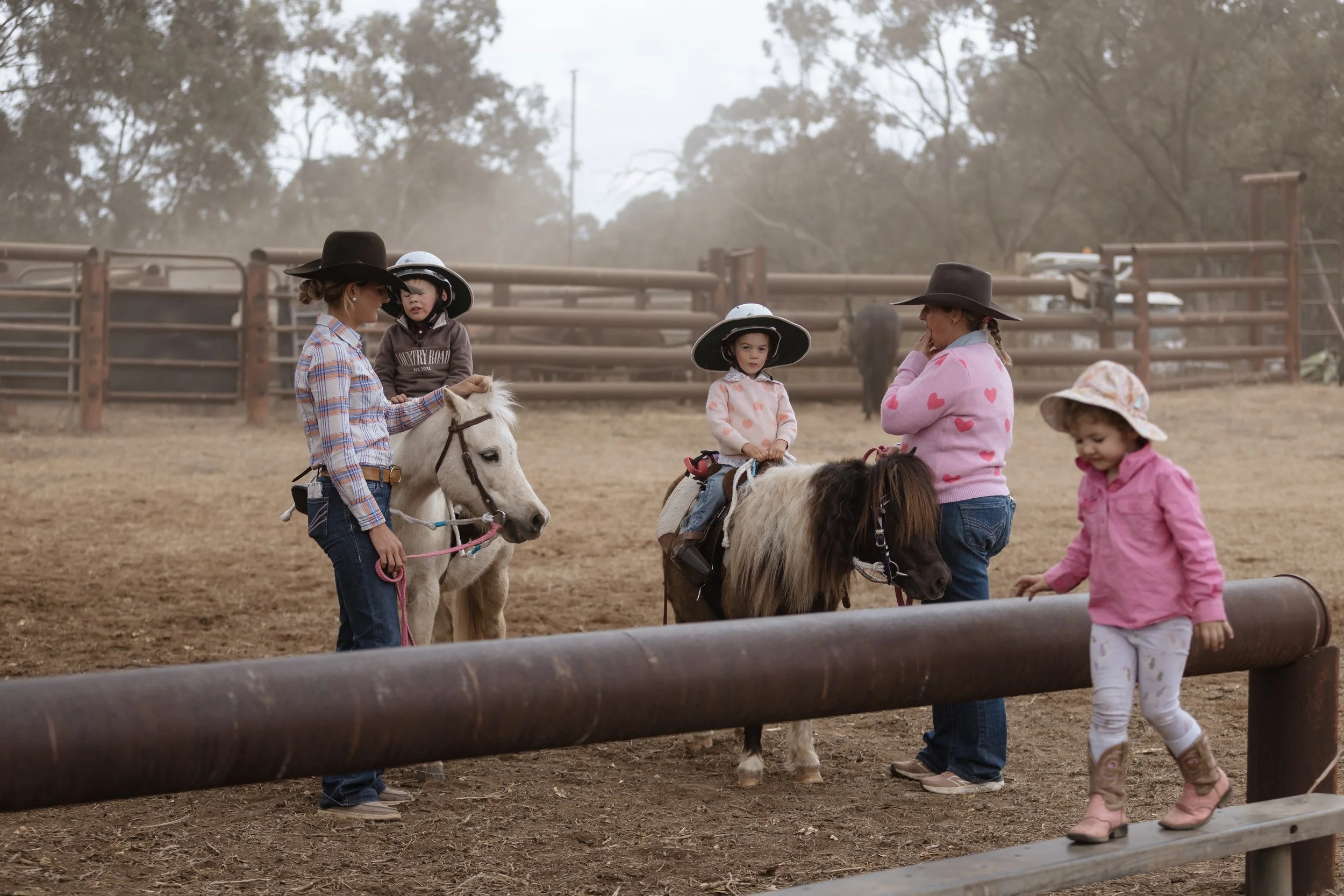 Children and adults riding ponies at a ranch with a wooden fence and trees in the background.