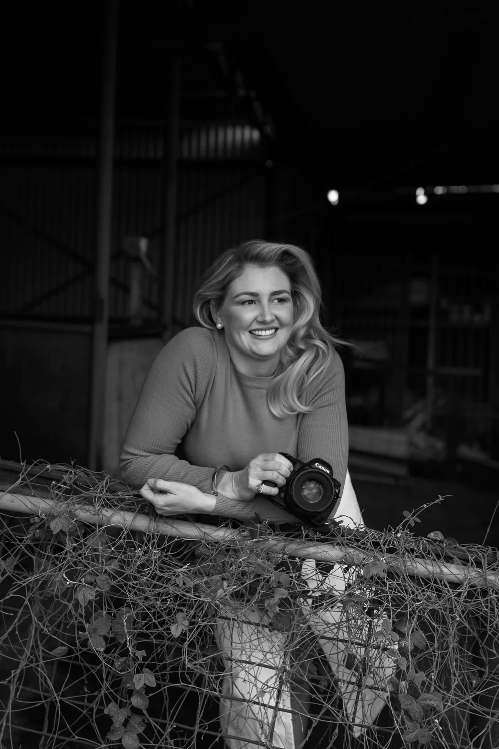 A smiling woman with wavy blonde hair wearing a long-sleeve top holding a Canon camera, leaning on a vine-covered fence in a dark, industrial-style setting.