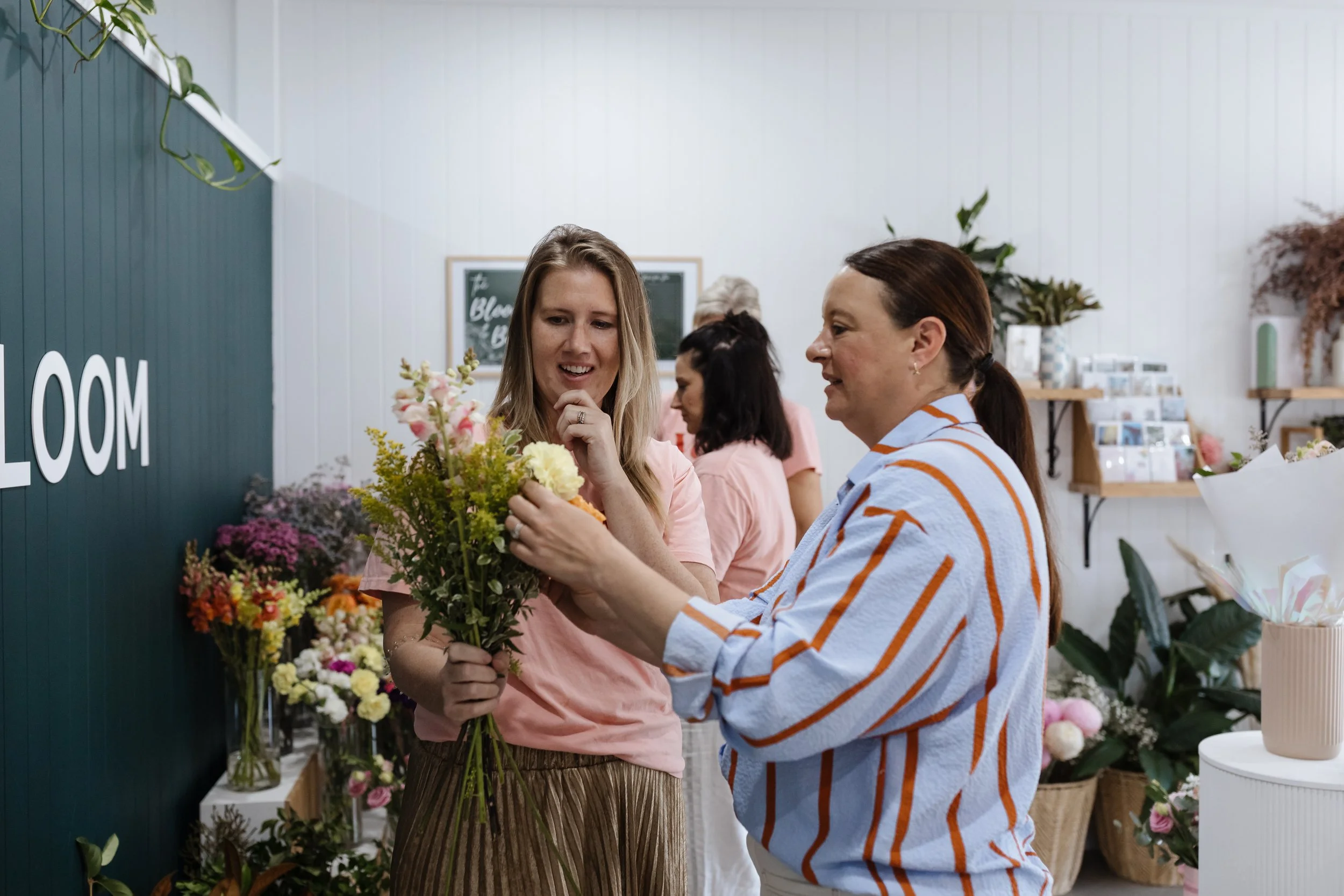 Two women shopping for flowers at a floral shop, one holding a bouquet of mixed flowers and the other examining it, with various plants and flowers displayed around them.
