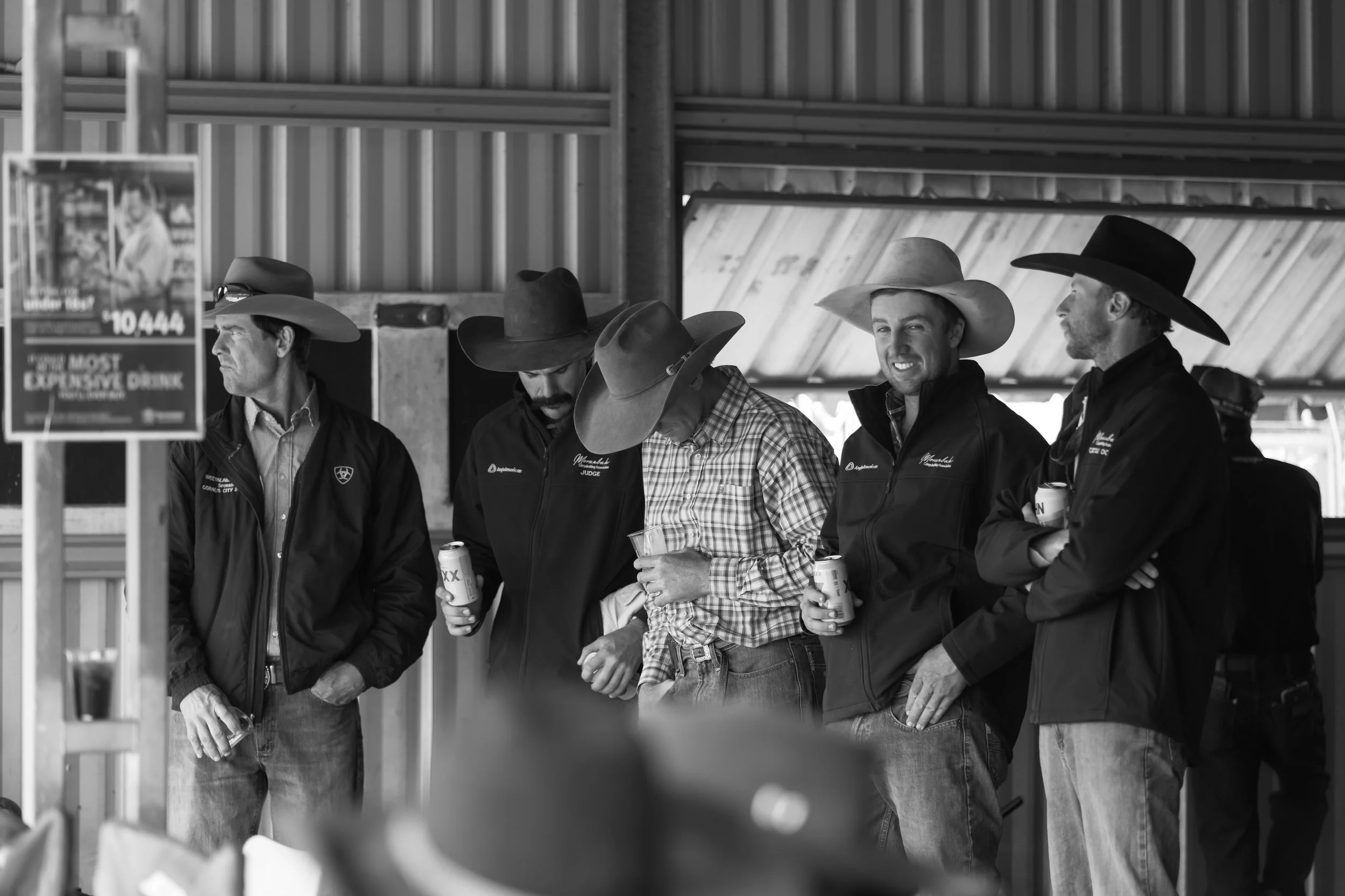 Black and white photo of five men wearing cowboy hats standing in line inside a wooden shelter. Some are holding cans, and they appear to be at an event or gathering.