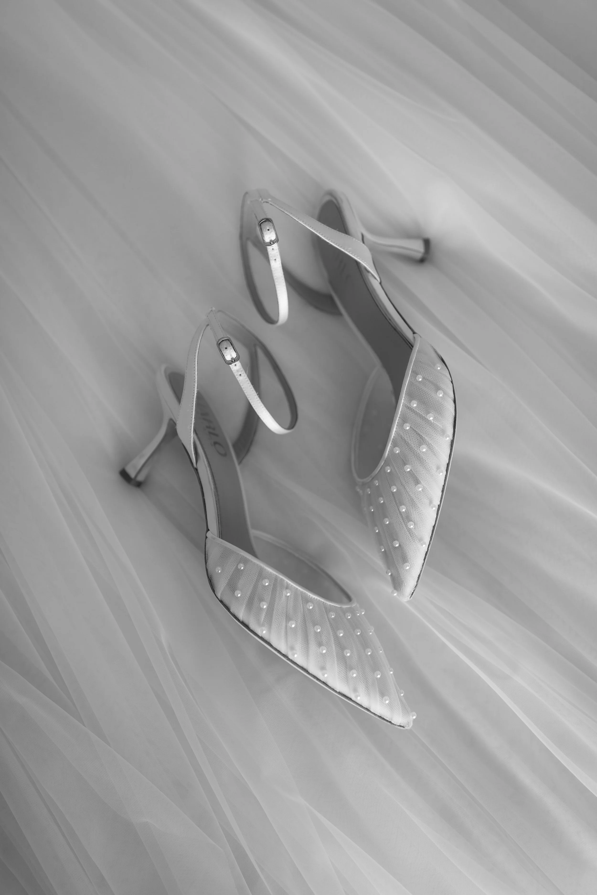 A pair of white high-heeled sandals with ankle straps and rhinestone embellishments, placed on a light-colored wooden surface.