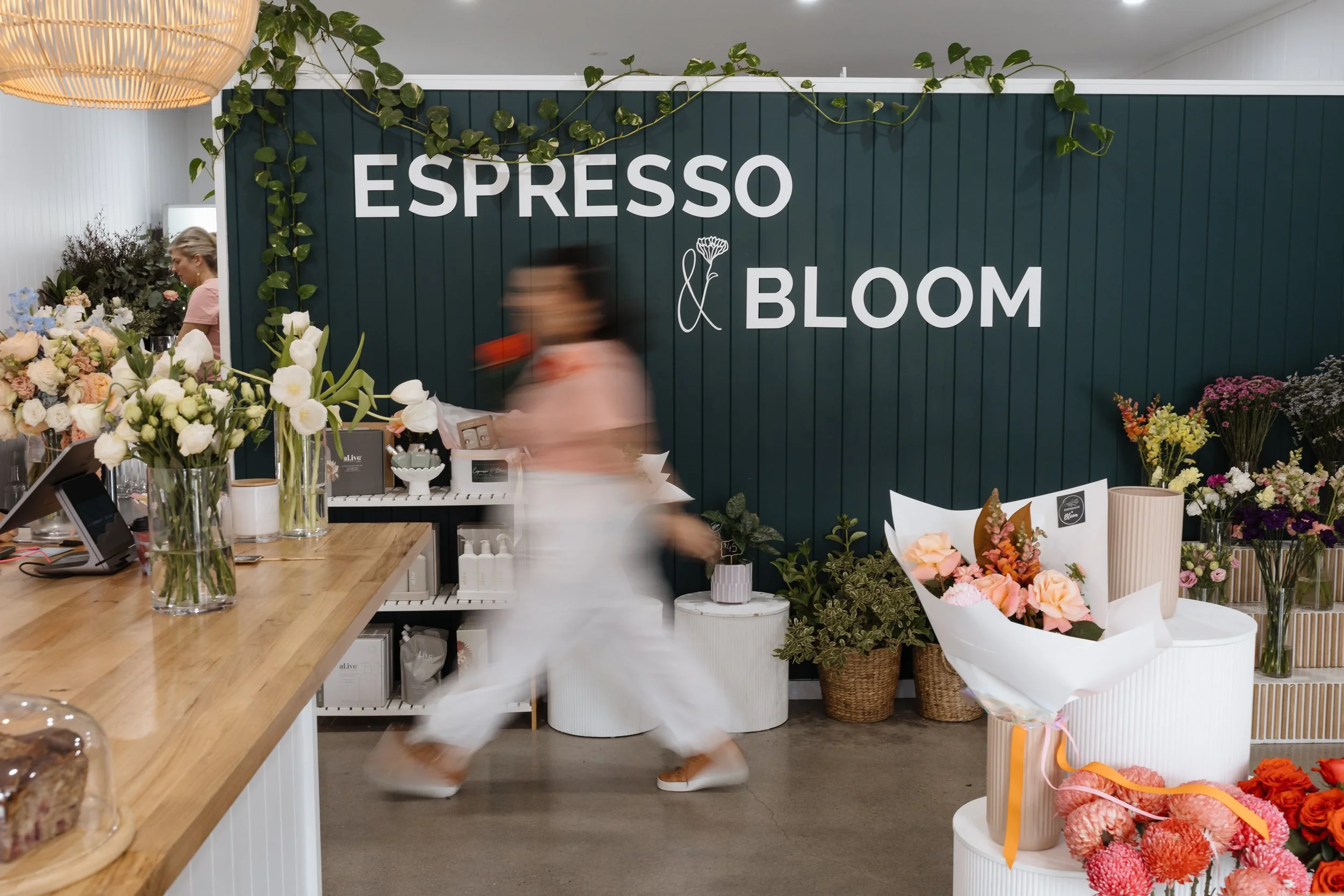 Interior of a flower shop with a dark green wall displaying the words 'ESPRESSO & BLOOM' in white letters, surrounded by various flowers and plants, with a woman walking past.