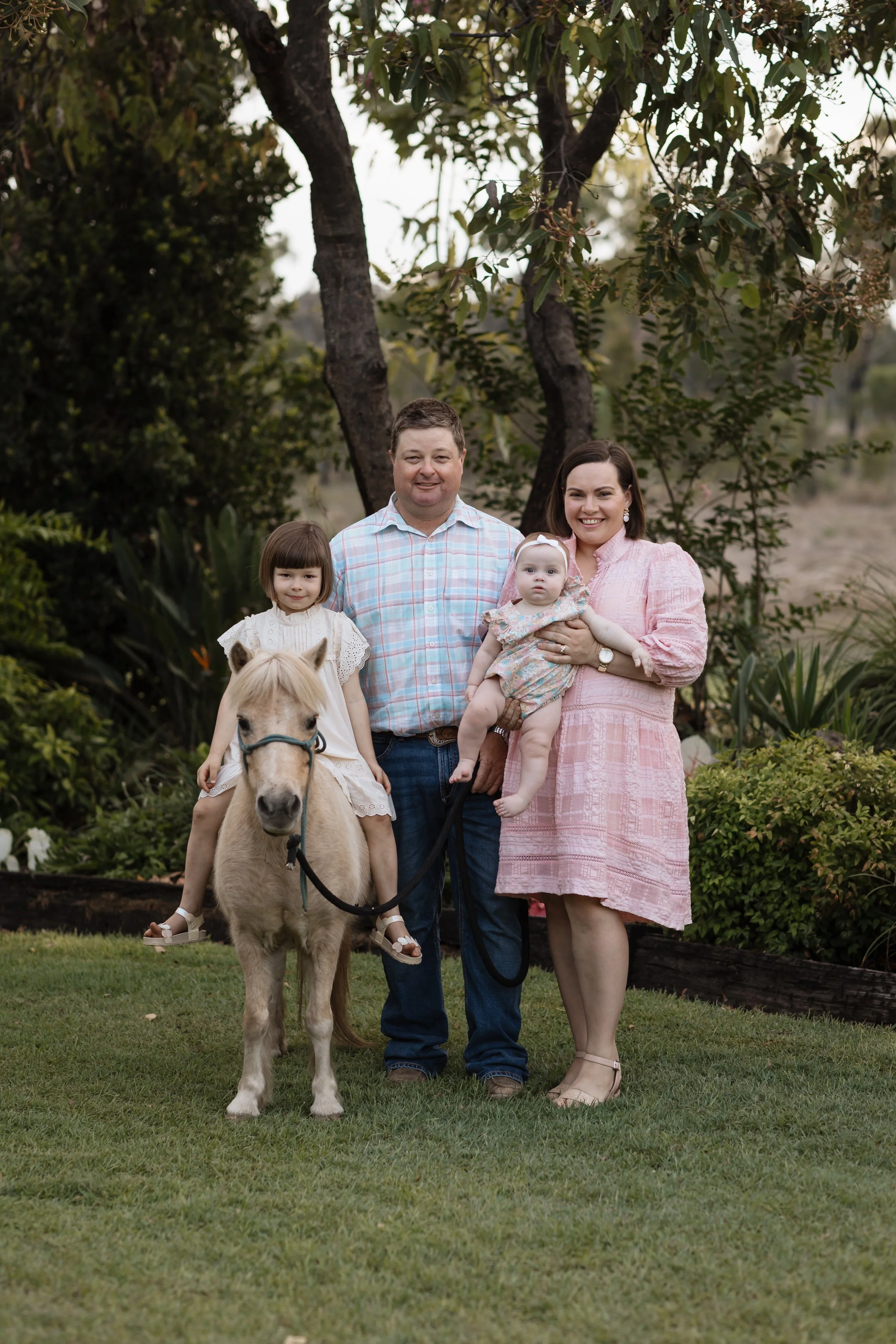 A family of four with a small dog and a pony standing on a grassy area near trees, smiling at the camera.