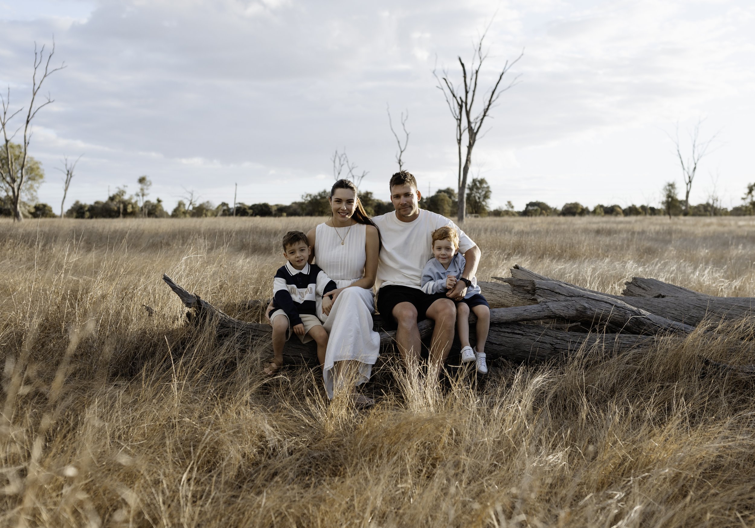 A family of four sitting on a fallen tree trunk in a field with tall, dry grass. The family includes a woman, a man, and two young boys. The background features scattered leafless trees and a cloudy sky.