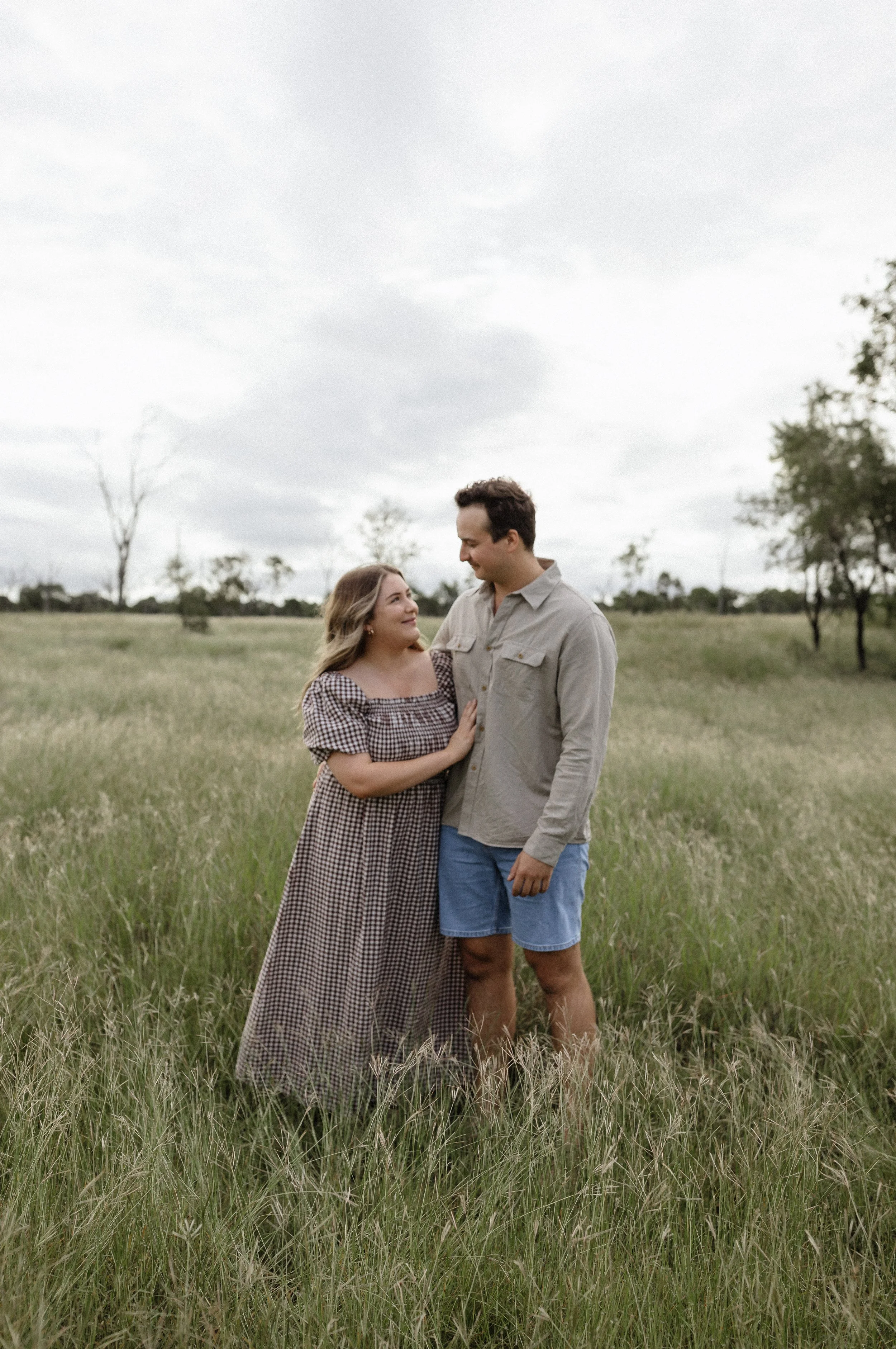 A young couple stands in a grassy field on a cloudy day, gazing at each other lovingly.