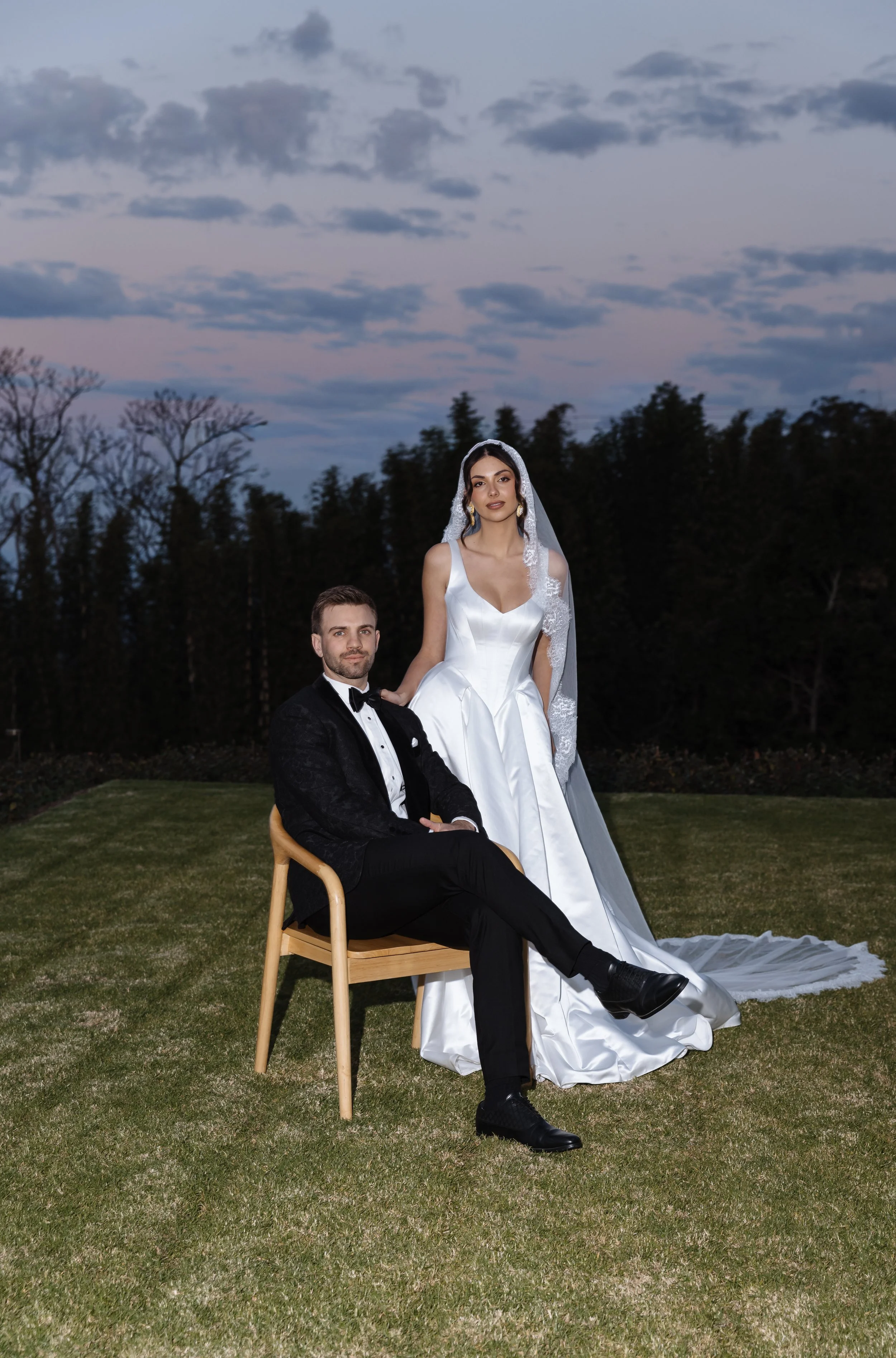 A man in a tuxedo sitting on a wooden chair with a woman in a wedding dress standing next to him outdoors during dusk.