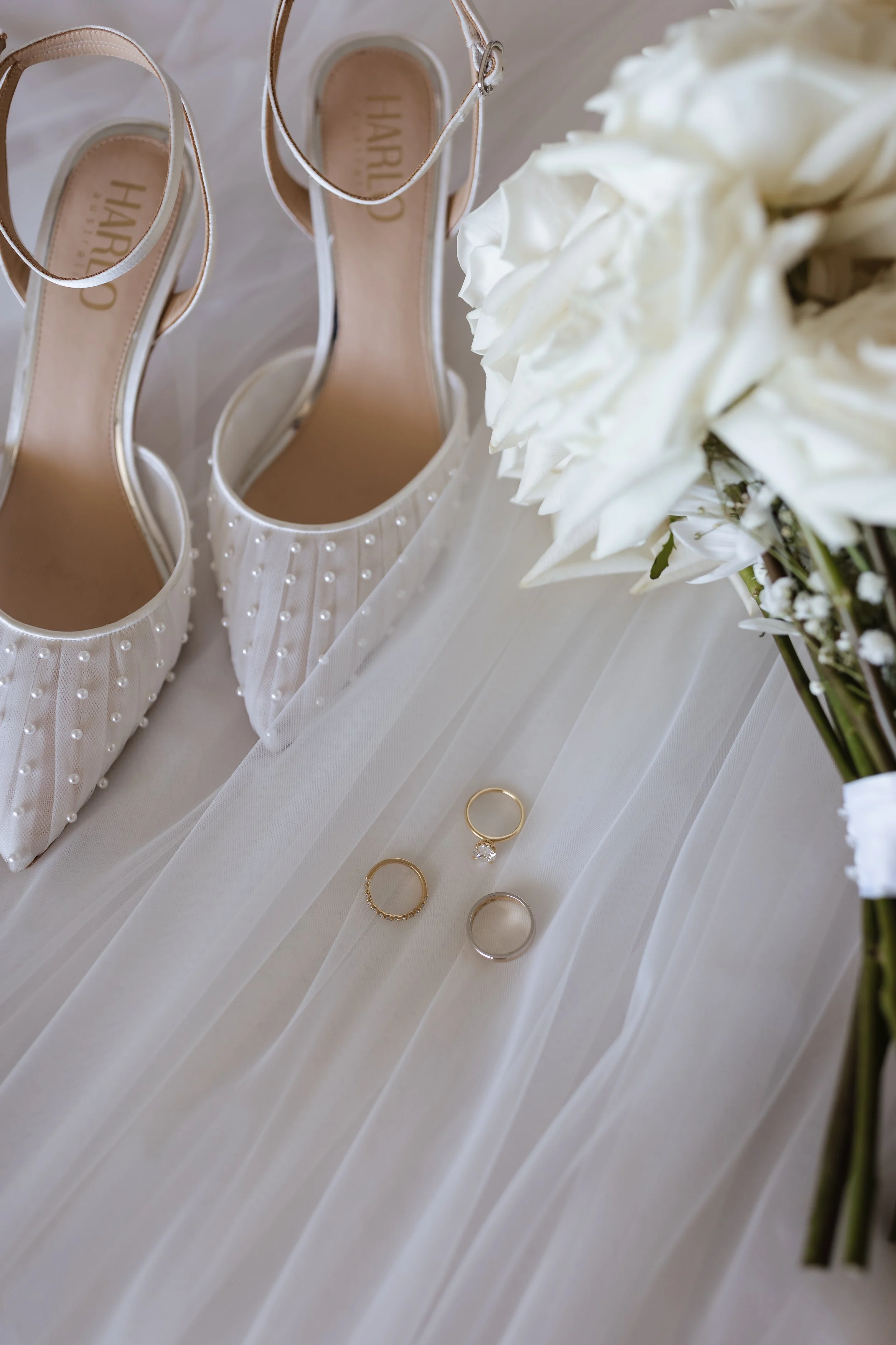 A pair of elegant white high-heeled shoes with pearl embellishments, a set of gold and silver rings, and a bouquet of white flowers arranged on a white fabric surface.