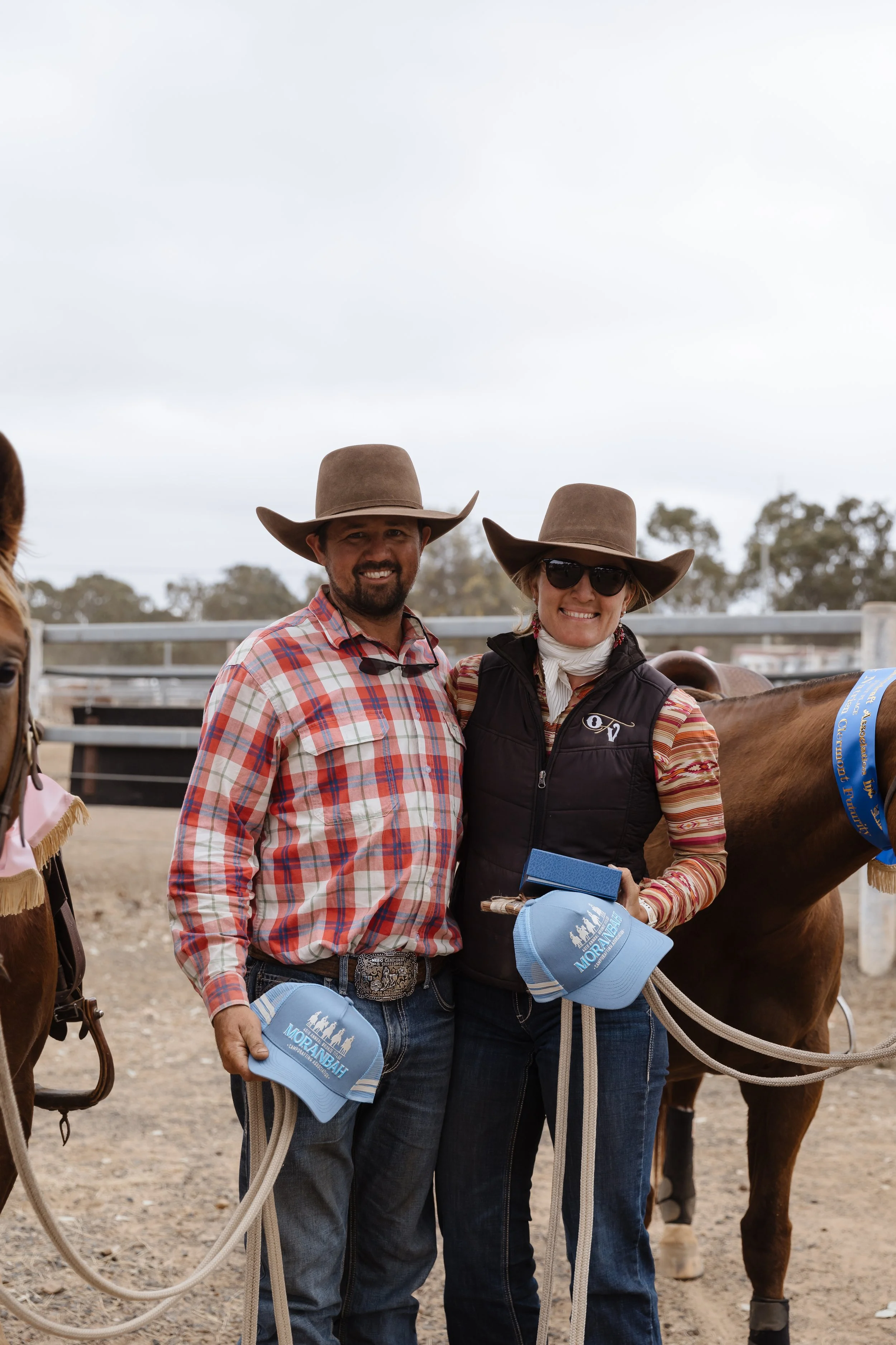 A man and a woman wearing cowboy hats and casual clothing, standing next to a horse at an outdoor ranch or farm. They are smiling and holding hats with a blue ribbon, indicating they might have participated in a rodeo or horse riding event.