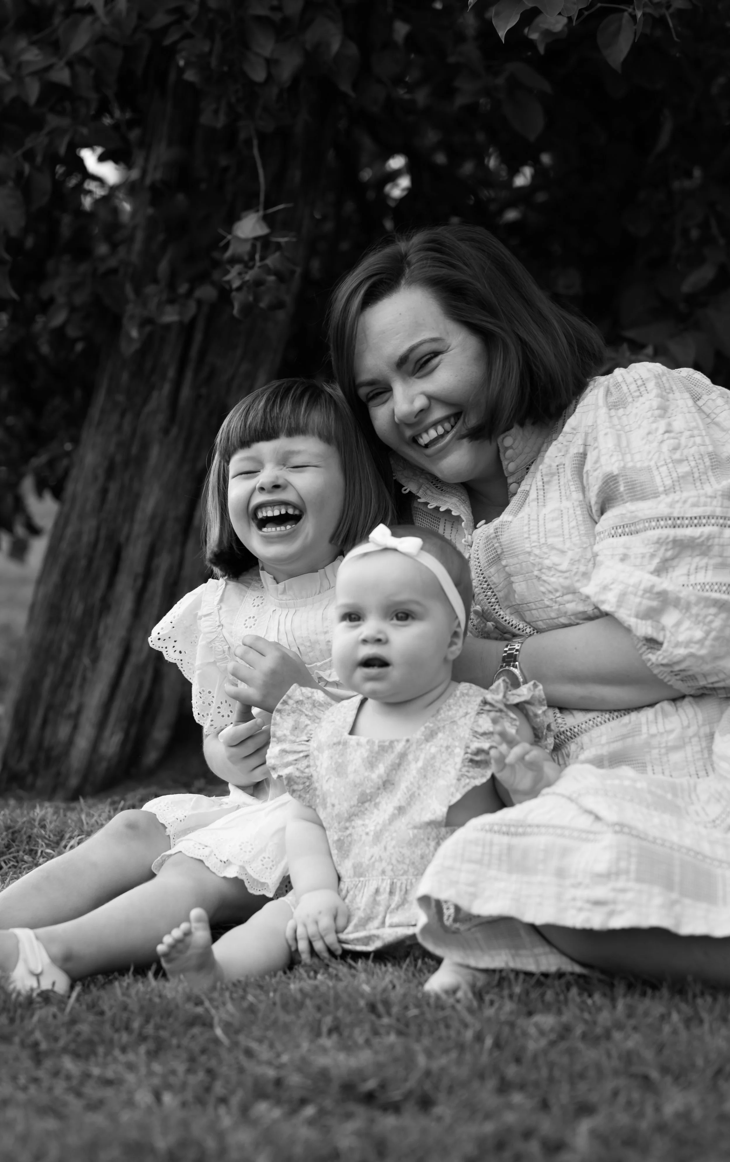 A woman and three young children sitting on the grass outdoors near a tree, all laughing and smiling.