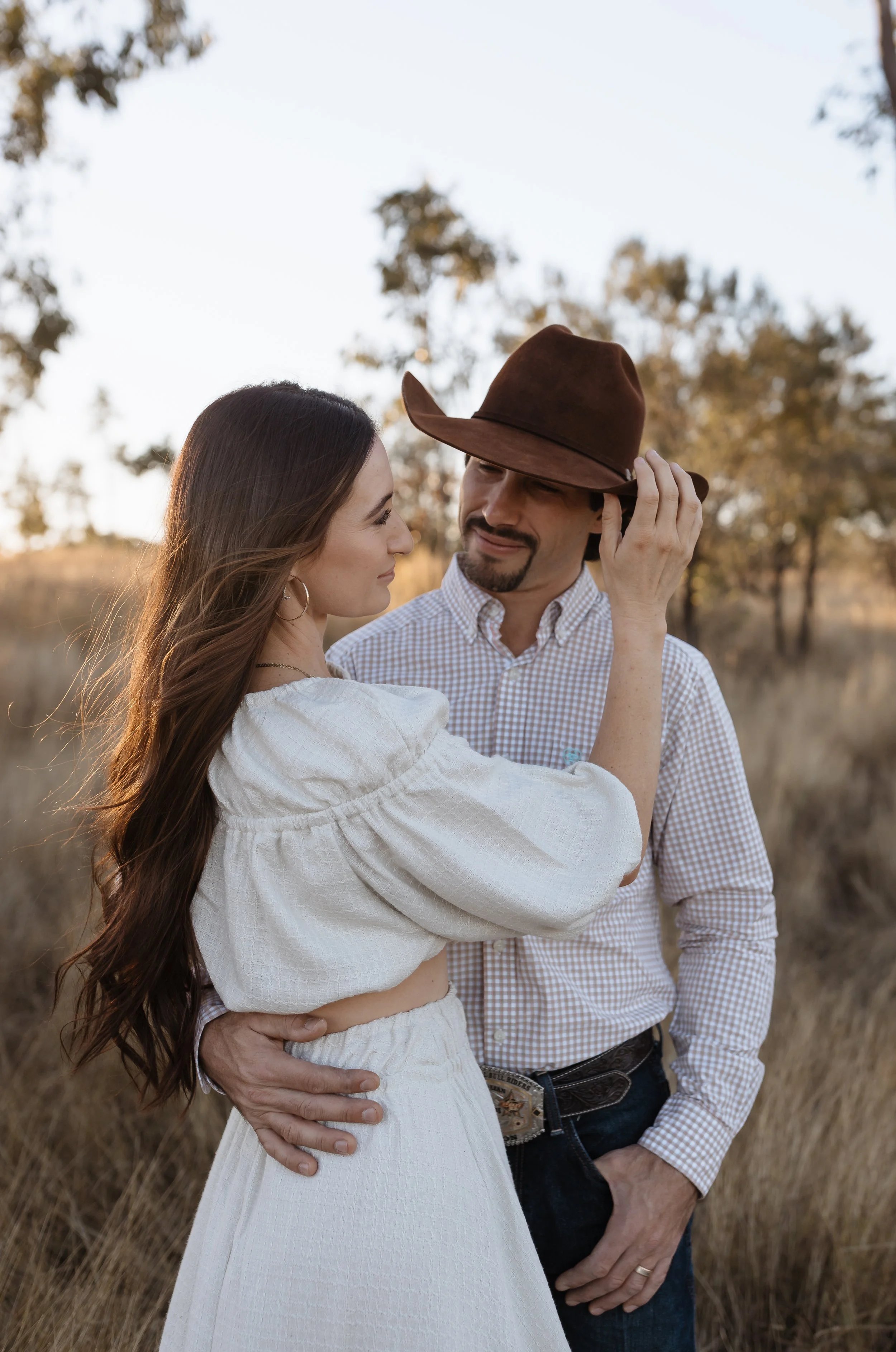 A couple stands close together outdoors in a field with trees in the background, woman touching the man's hat as they look at each other warmly.