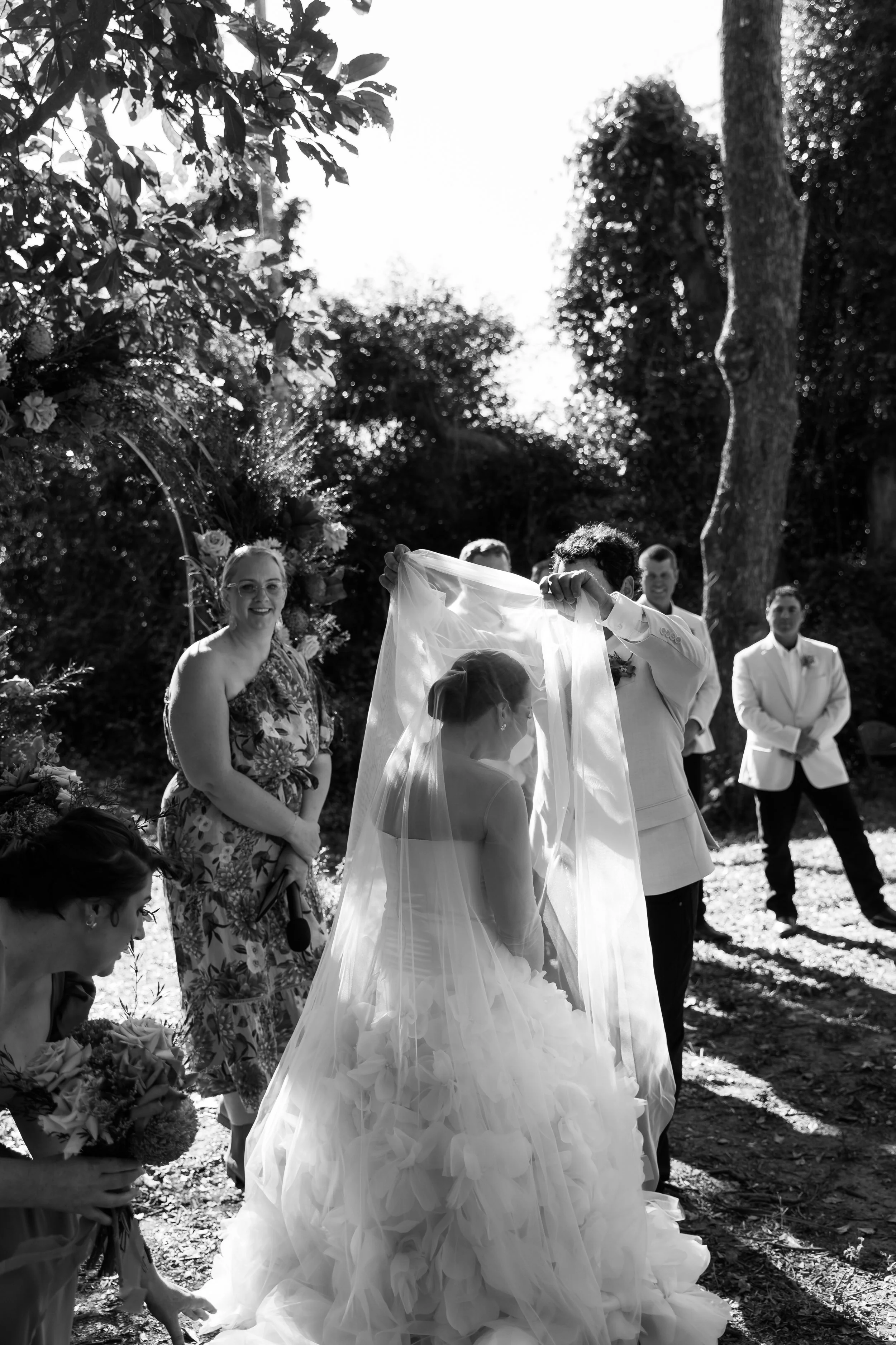 A bride in a wedding dress with a long, voluminous skirt and veil stands outdoors during a wedding ceremony. She is surrounded by her wedding party and guests, with some people holding bouquets of flowers. The ceremony takes place under trees with su
