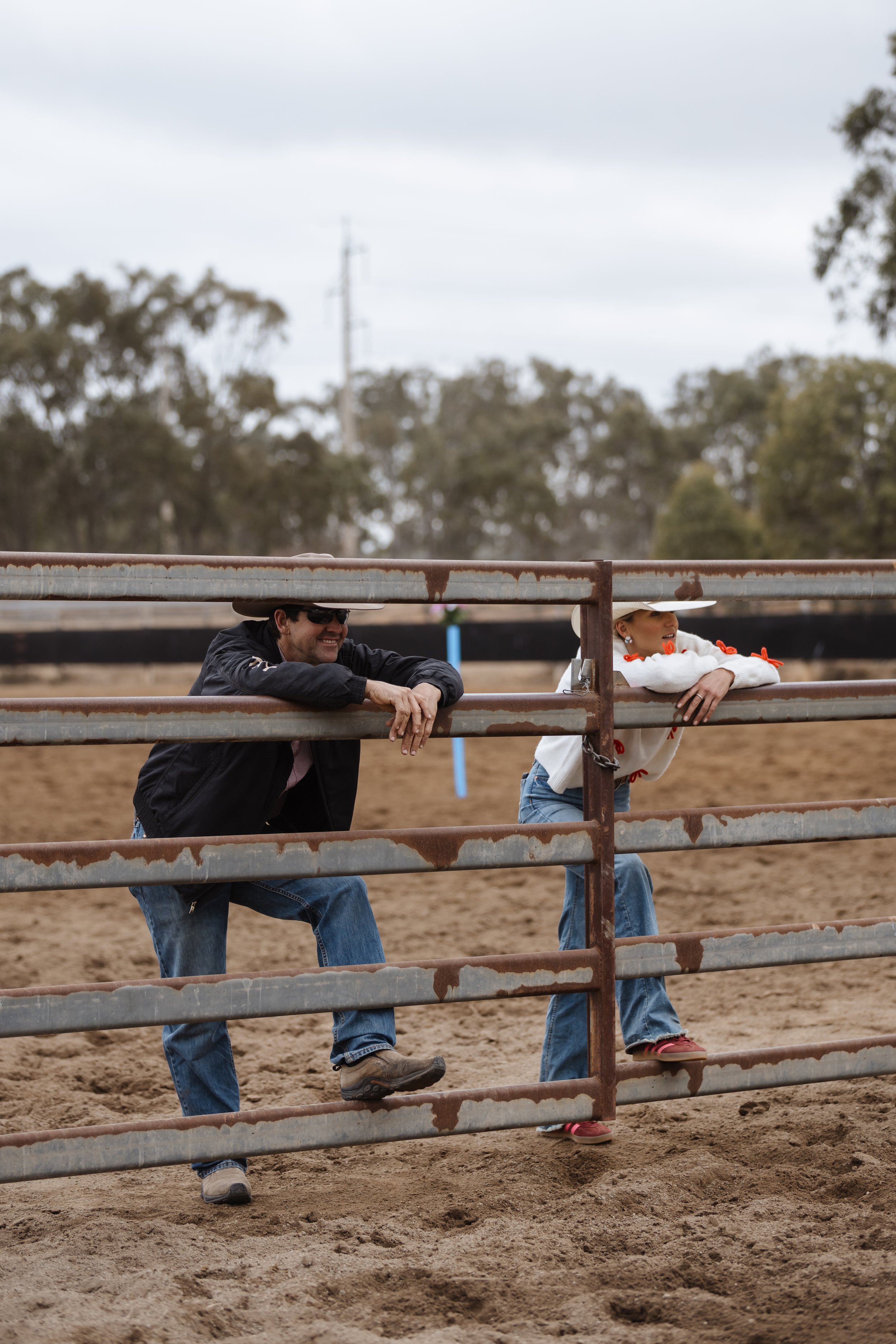 Two people leaning on a rusty metal fence at a rodeo or similar event, with a dirt ground and trees in the background.