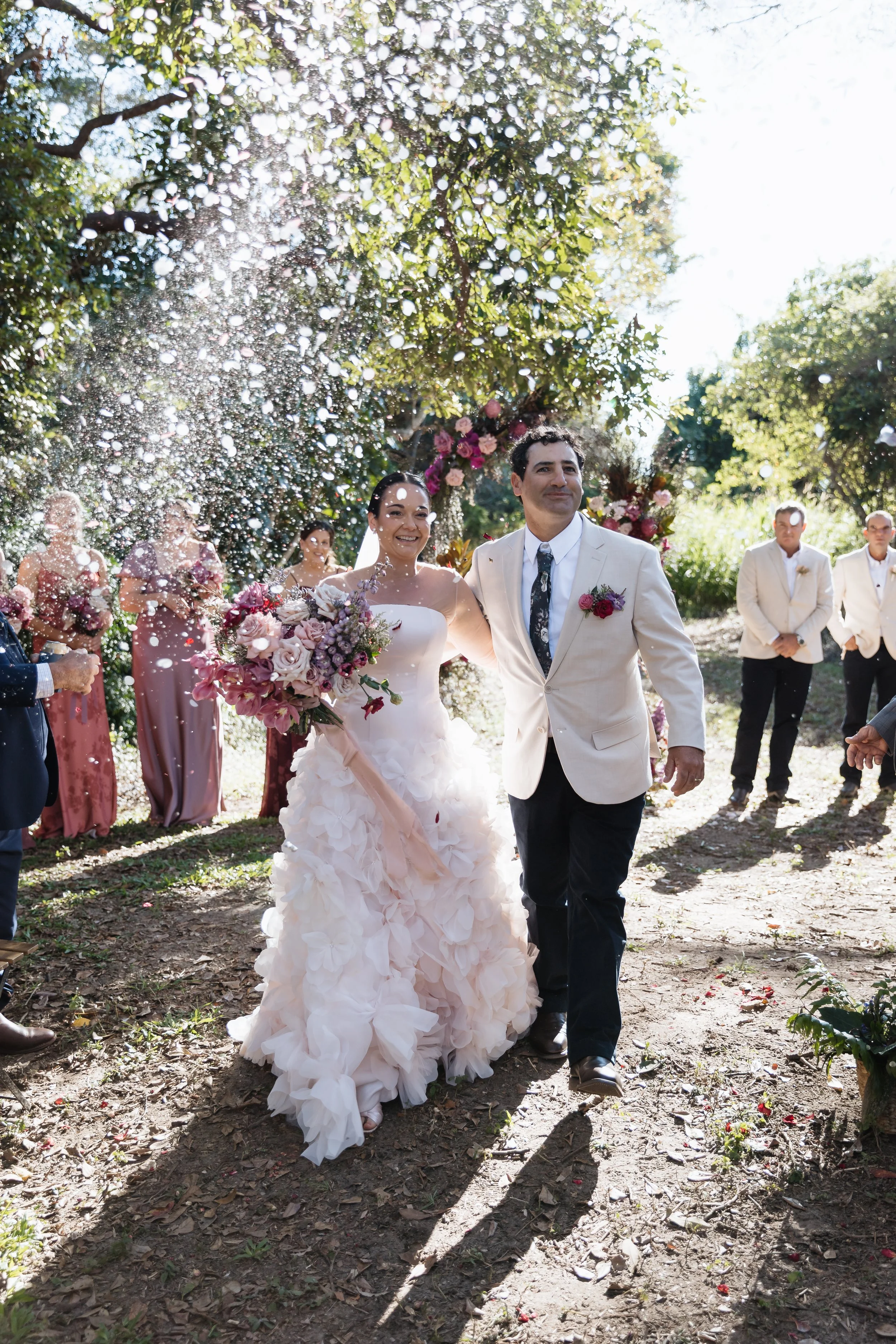 A newly married couple, the bride in a white wedding gown holding a bouquet of pink and purple flowers, and the groom in a beige suit with a floral tie, walking outdoors under a floral arch with wedding guests and groomsmen in the background. The sce