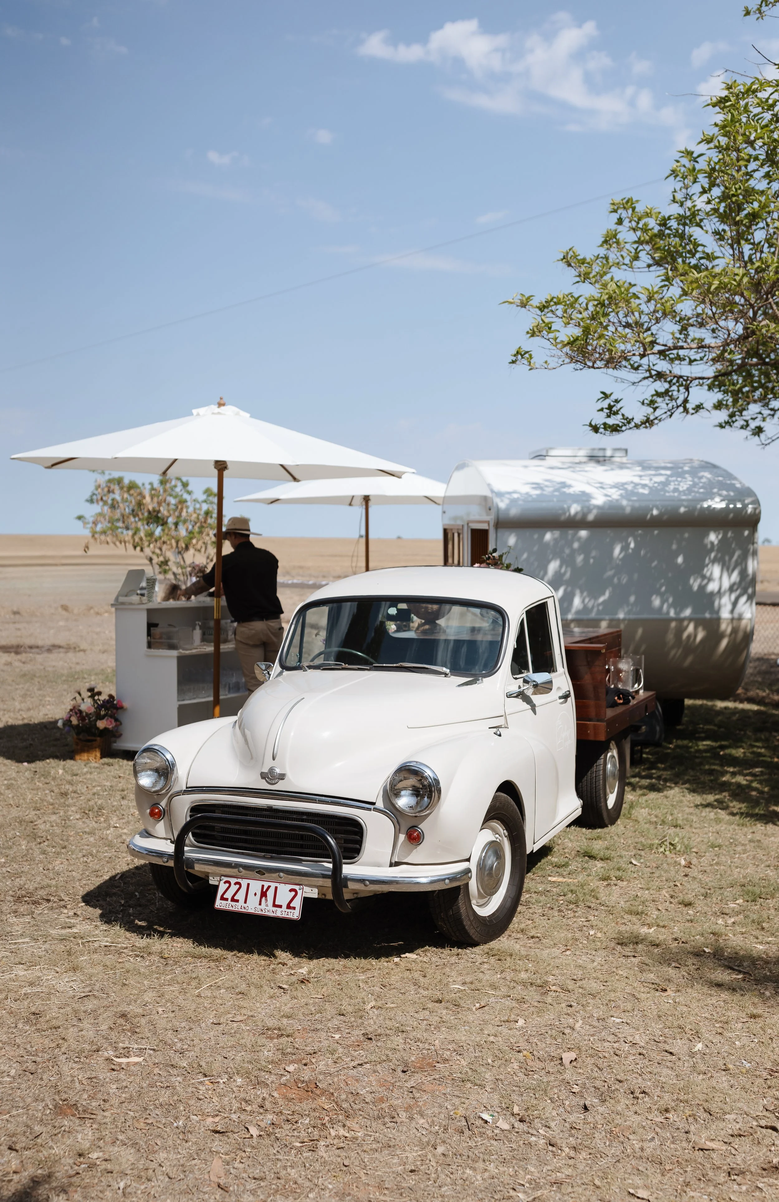A vintage white car with Queensland license plates parked on a grassy field, with a small trailer hitched to it. In the background, there are white umbrellas, a man in dark clothing and a hat serving at a white table, and a tree with green leaves. The sky is blue with a few scattered clouds.