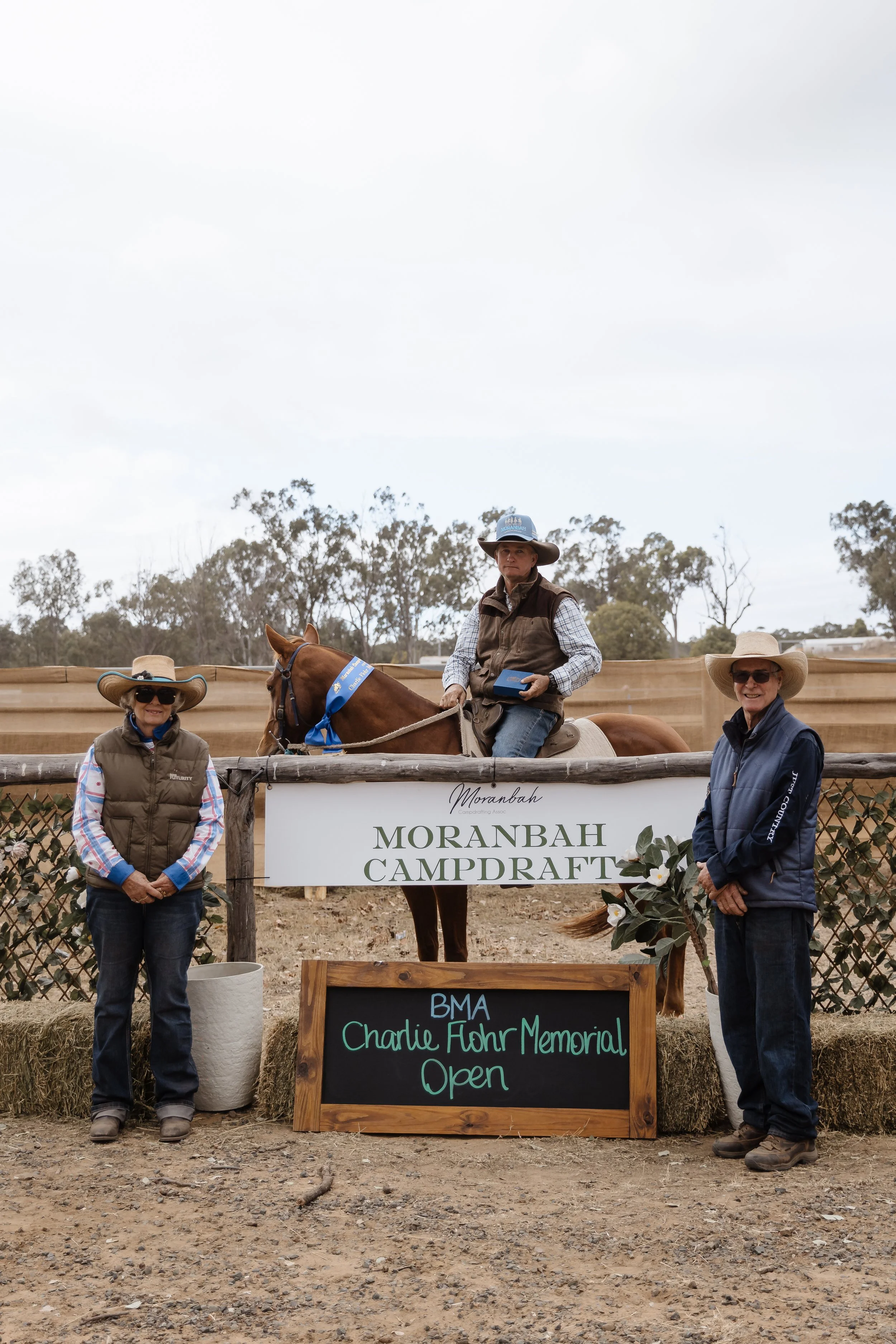 Group of people at Morambah Campdraft event, with a woman and a man standing on either side, and a person on horseback in the middle, holding a ribbon. There is a sign for the Charlie Fohr Memorial Open, and decorative flowers in the foreground.
