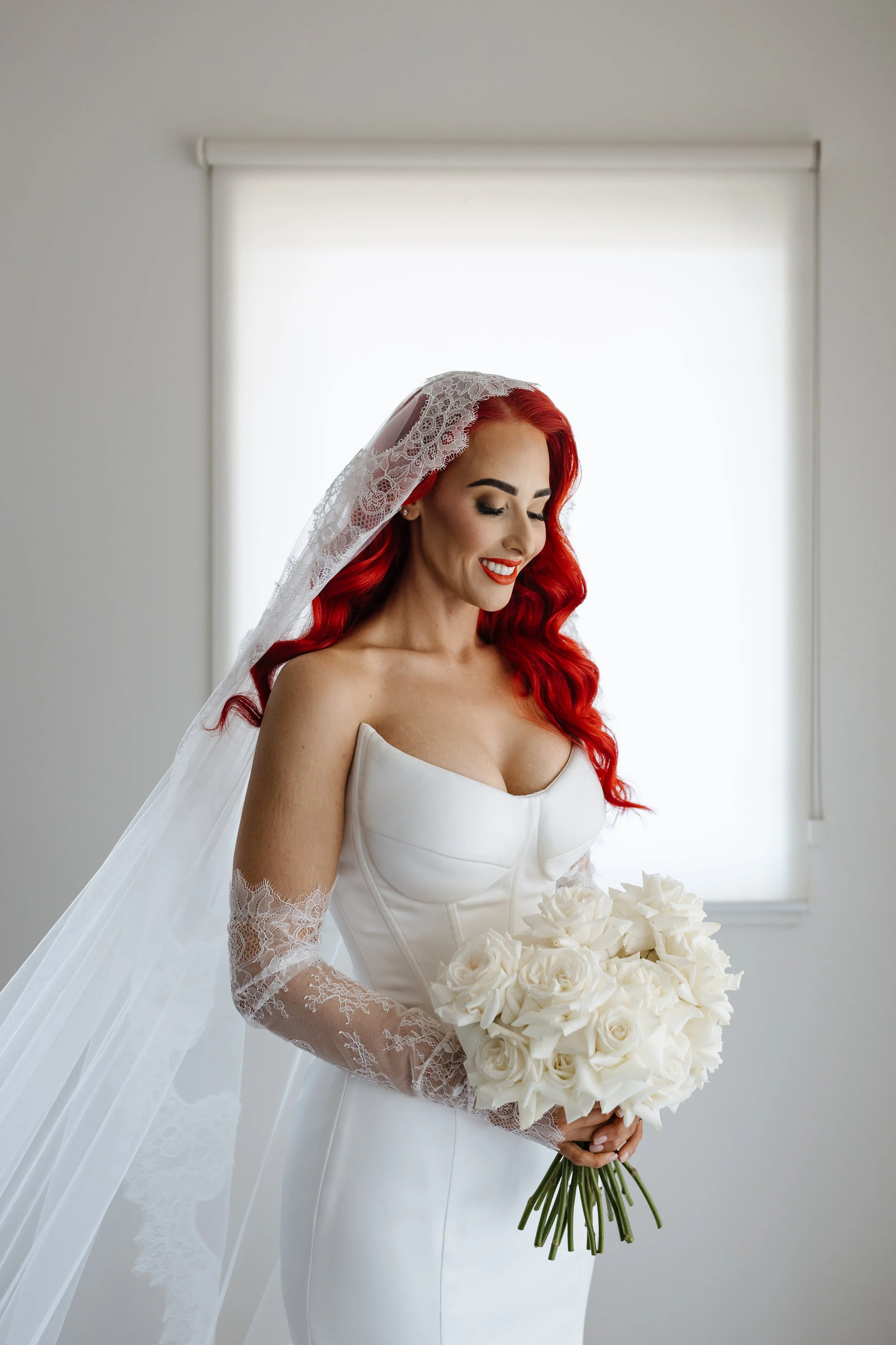 A woman with long red hair, dressed in a white wedding gown with satin and lace details, holding a bouquet of white roses, standing in front of a window with white blinds.