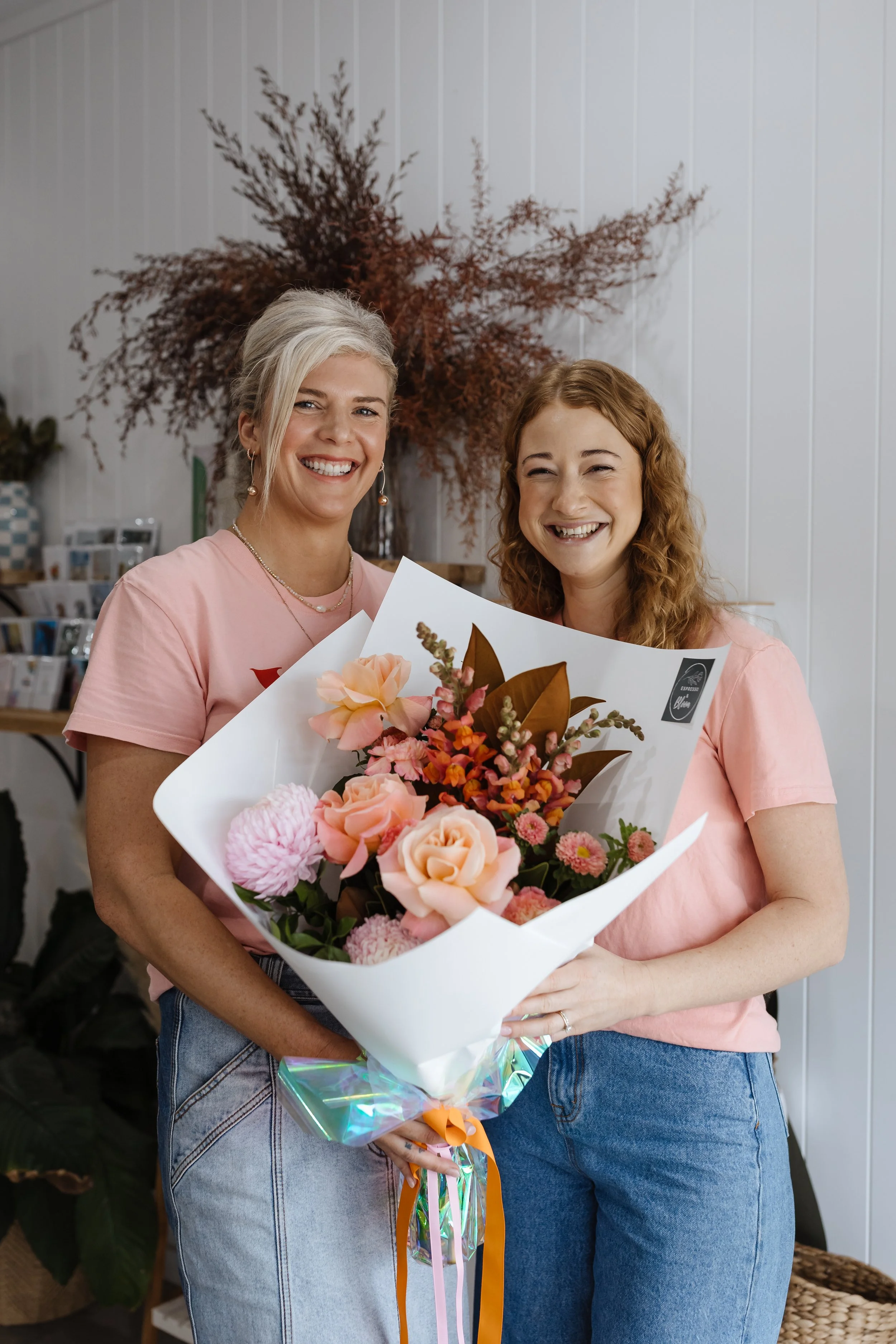 Two women smiling and holding a bouquet of pink and peach flowers wrapped in white paper. One woman has blonde hair and is wearing a pink t-shirt and blue jeans, and the other has red hair and is also wearing a pink t-shirt and blue jeans. They are s