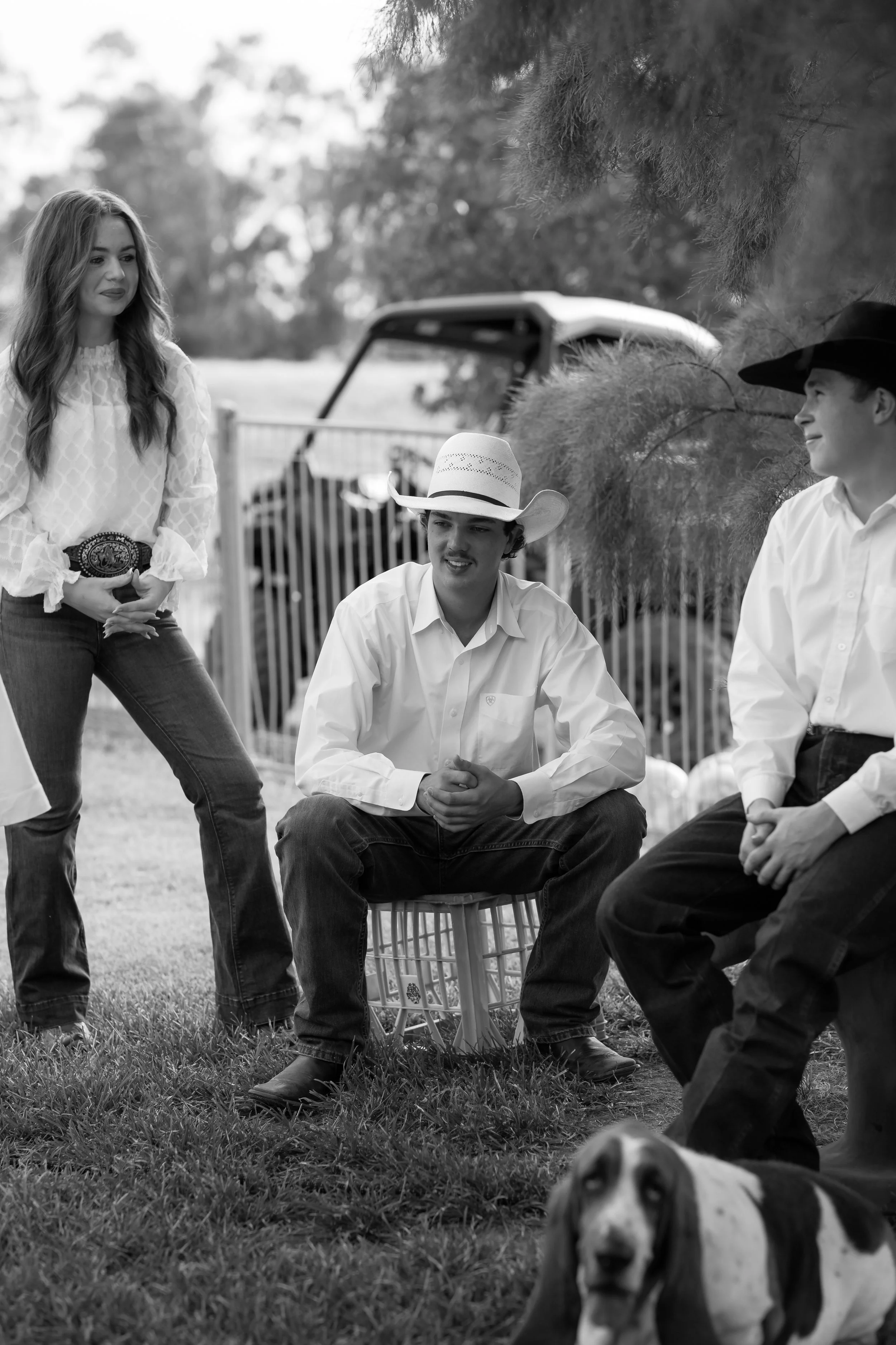 Four people, two men and one woman, are outdoors, with one man wearing a cowboy hat. The woman is standing, and the two men are sitting; one is in a cowboy hat, and the other is in a black hat. There is a dog lying on the grass in the foreground. Tre